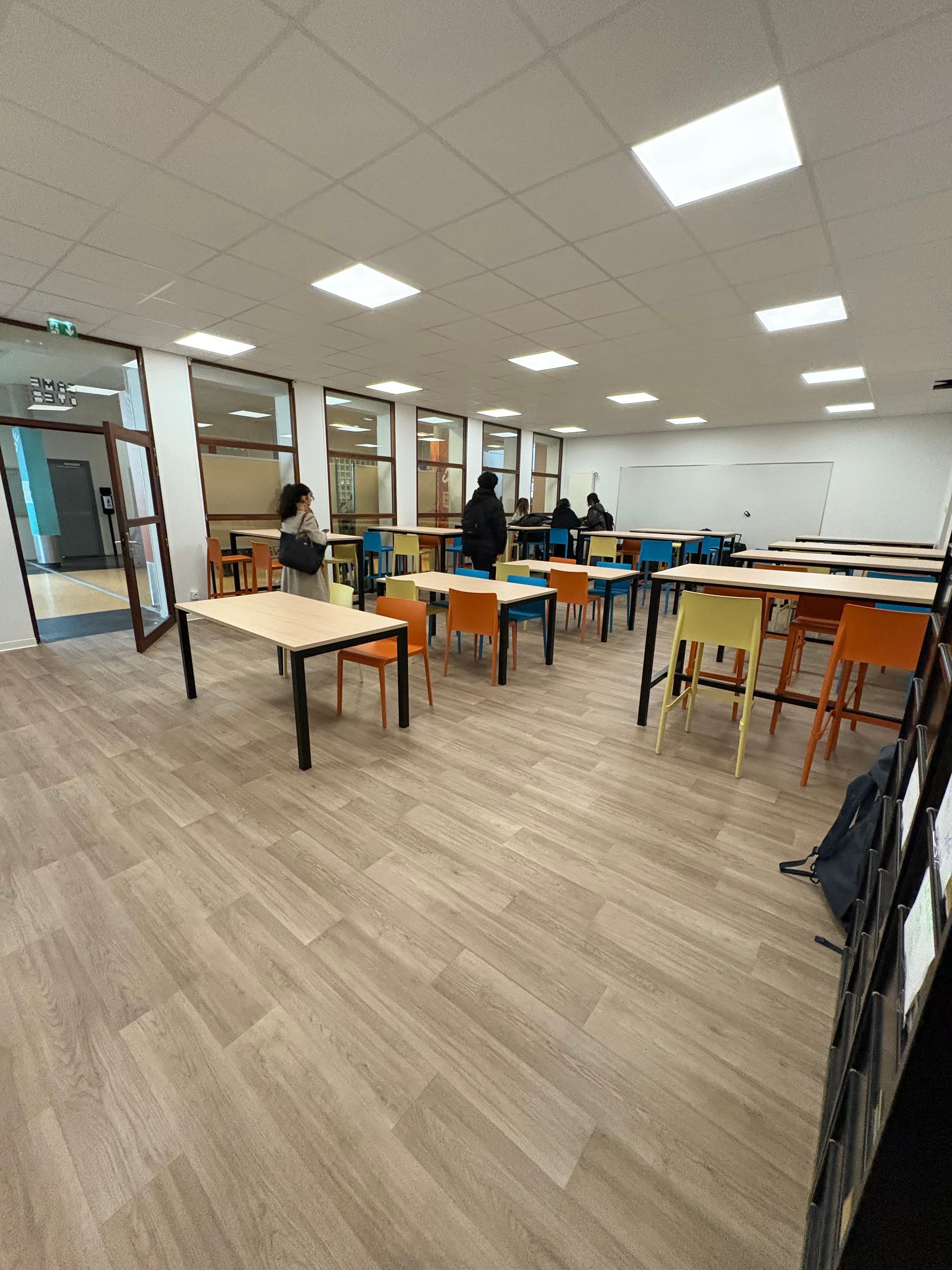 Un espace d'étude ou une bibliothèque moderne et lumineuse, avec des rangées de tables en bois clair, des chaises colorées et des murs vitrés.