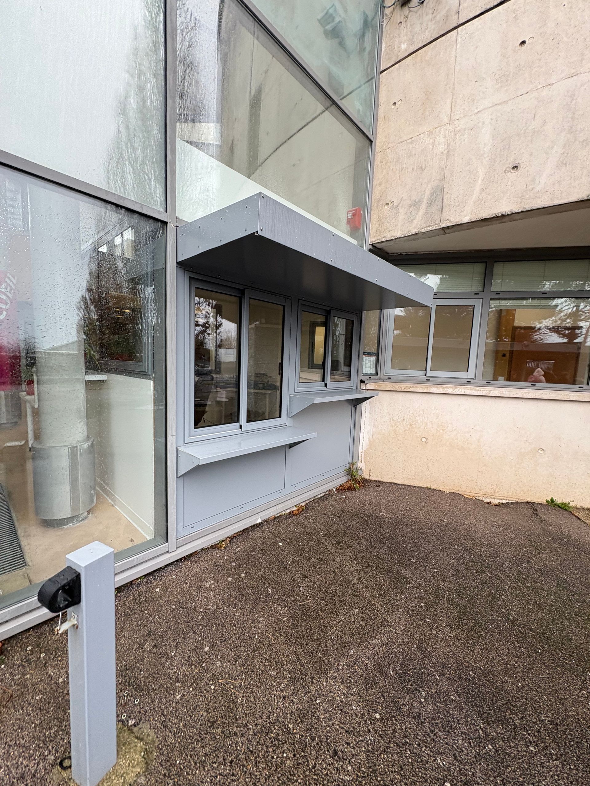 Une fenêtre de service grise et couverte, construite sur le mur extérieur d'un bâtiment situé à côté d'une zone gravillonnée.