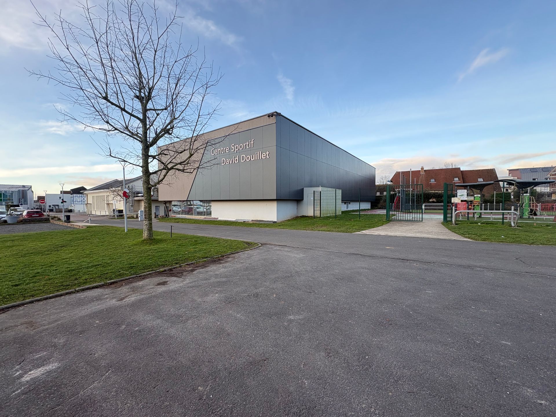 Un bâtiment commercial gris moderne se dresse à côté d'un arbre sans feuilles sous un ciel bleu, avec un parking pavé au premier plan.