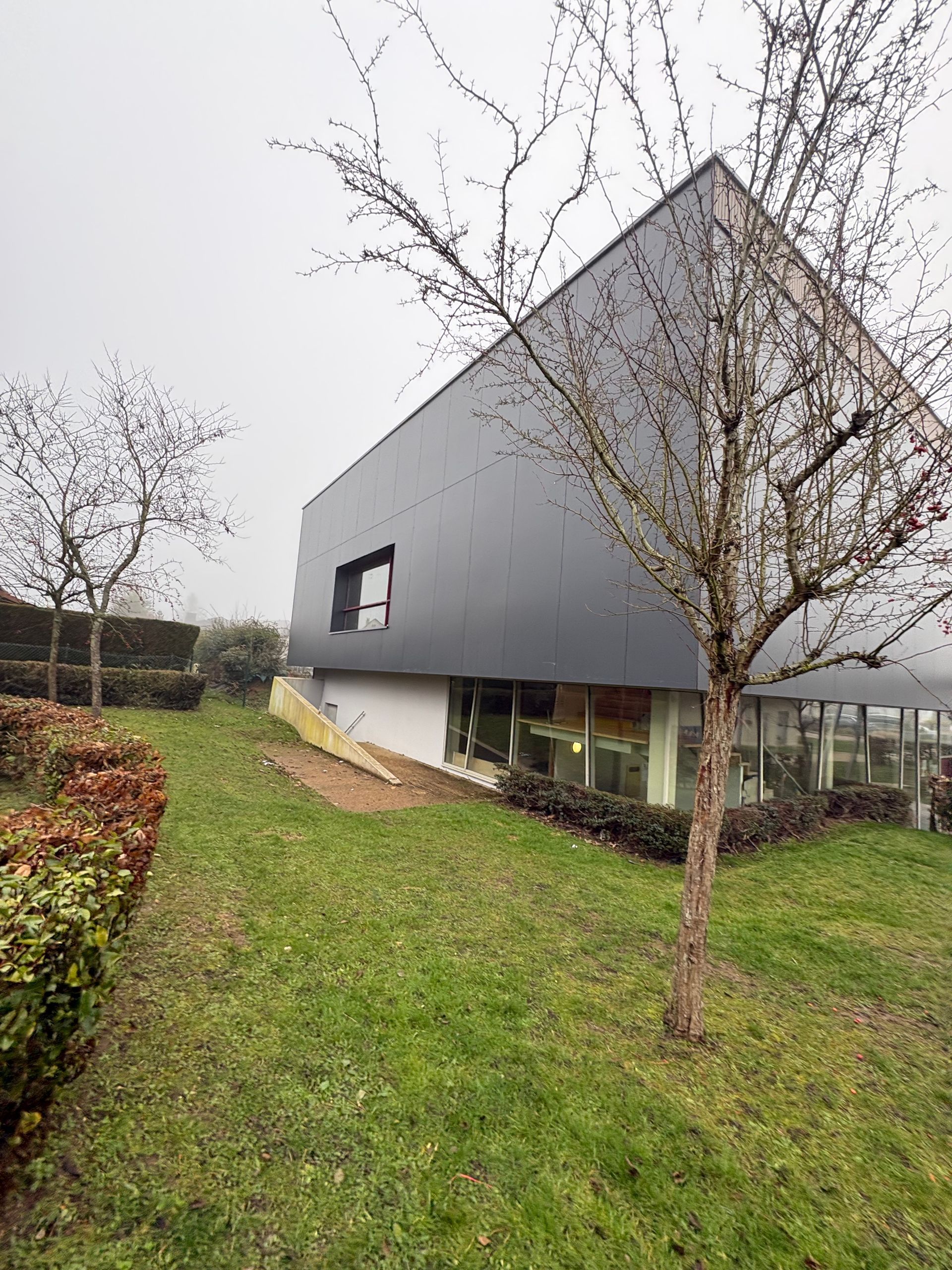 Une maison moderne, avec un étage supérieur en métal sombre et un rez-de-chaussée blanc, se dresse dans une cour verdoyante aux arbres dénudés, par une journée nuageuse.