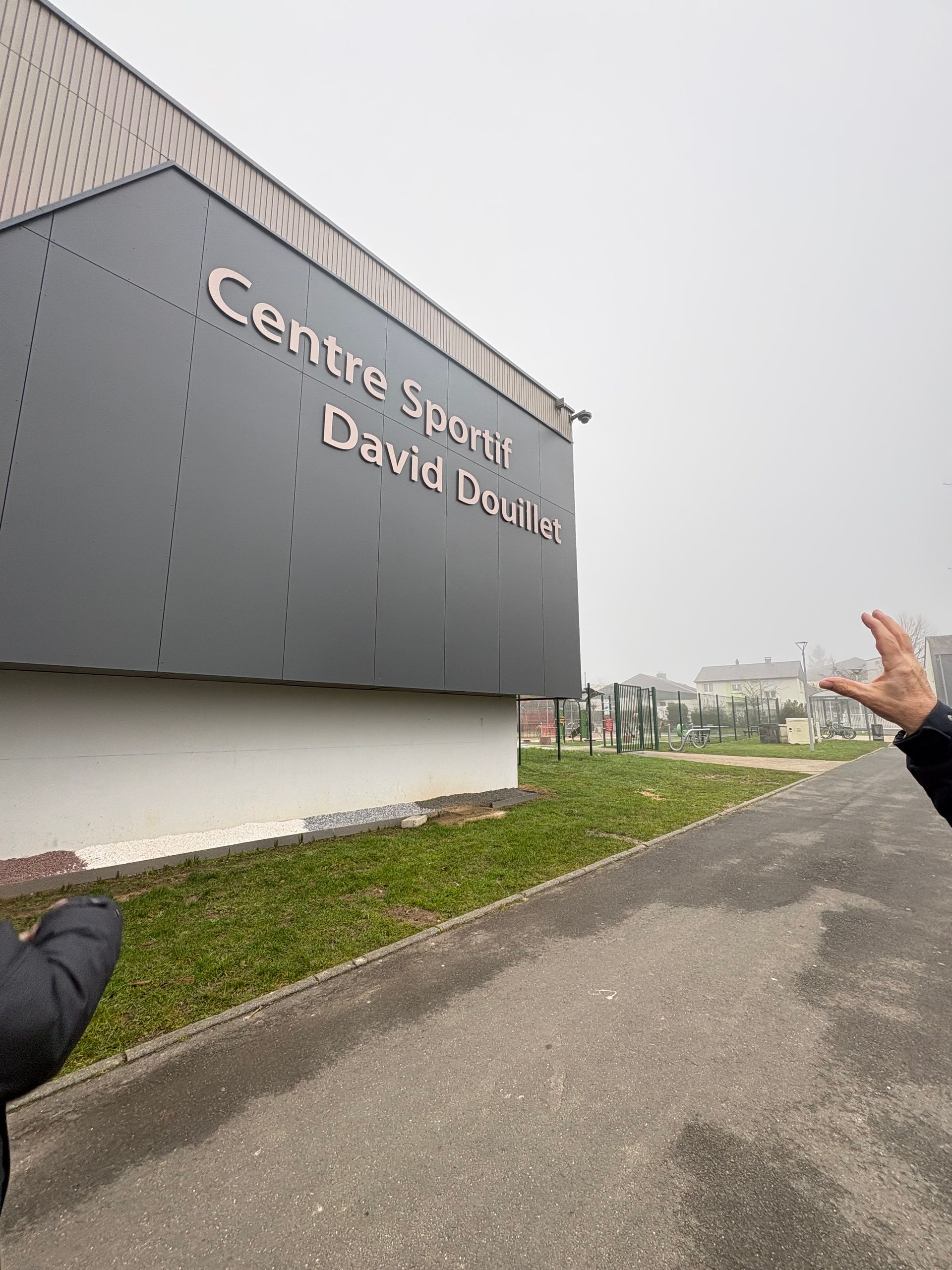 Une personne fait un geste de la main en direction du bâtiment du « Centre Sportif David Douillet » sous un ciel brumeux et couvert.