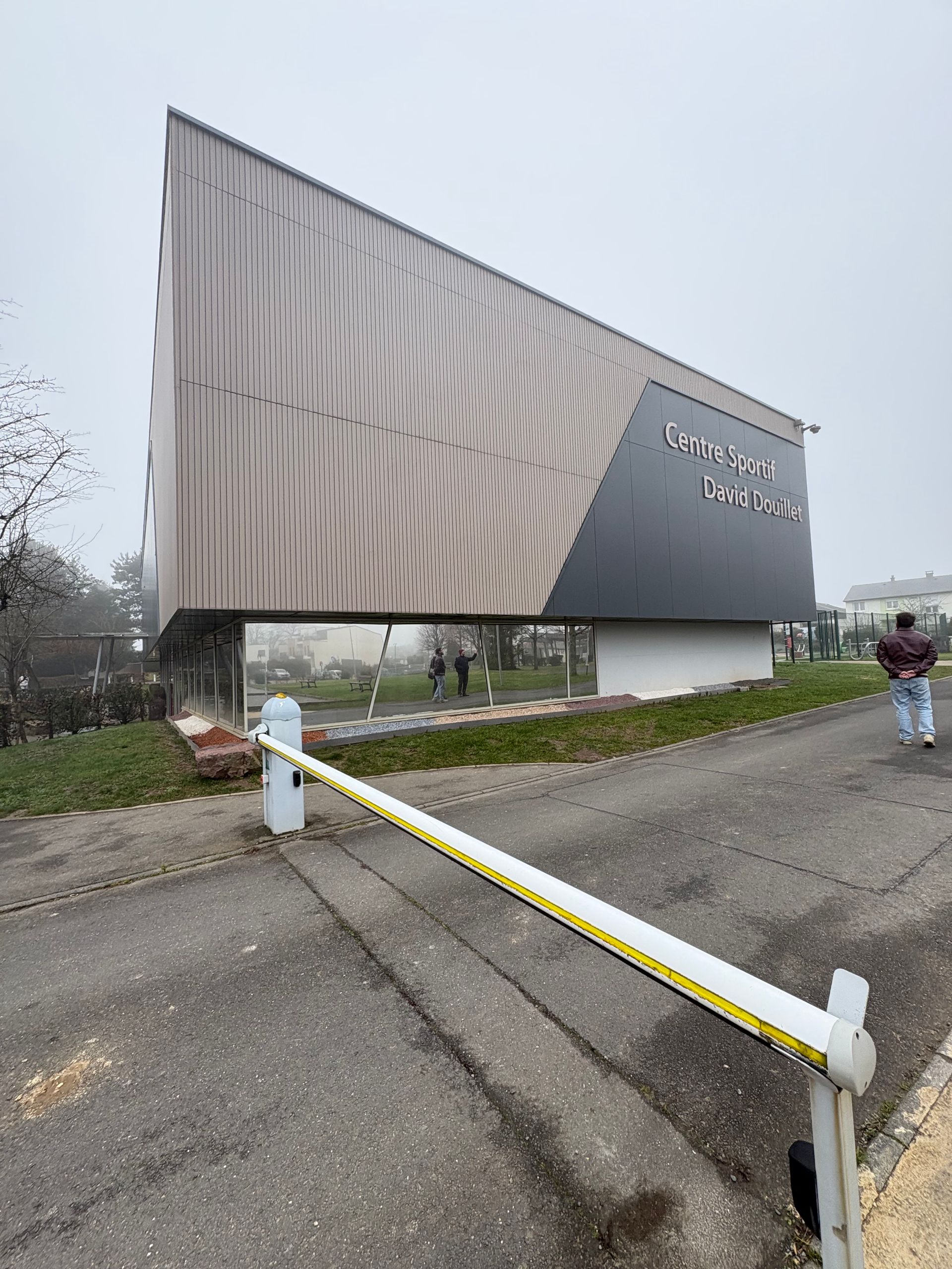 Un bâtiment moderne aux lignes angulaires, revêtu d'un bardage à motifs de bois, surplombe un portail près d'une route asphaltée par une journée brumeuse.