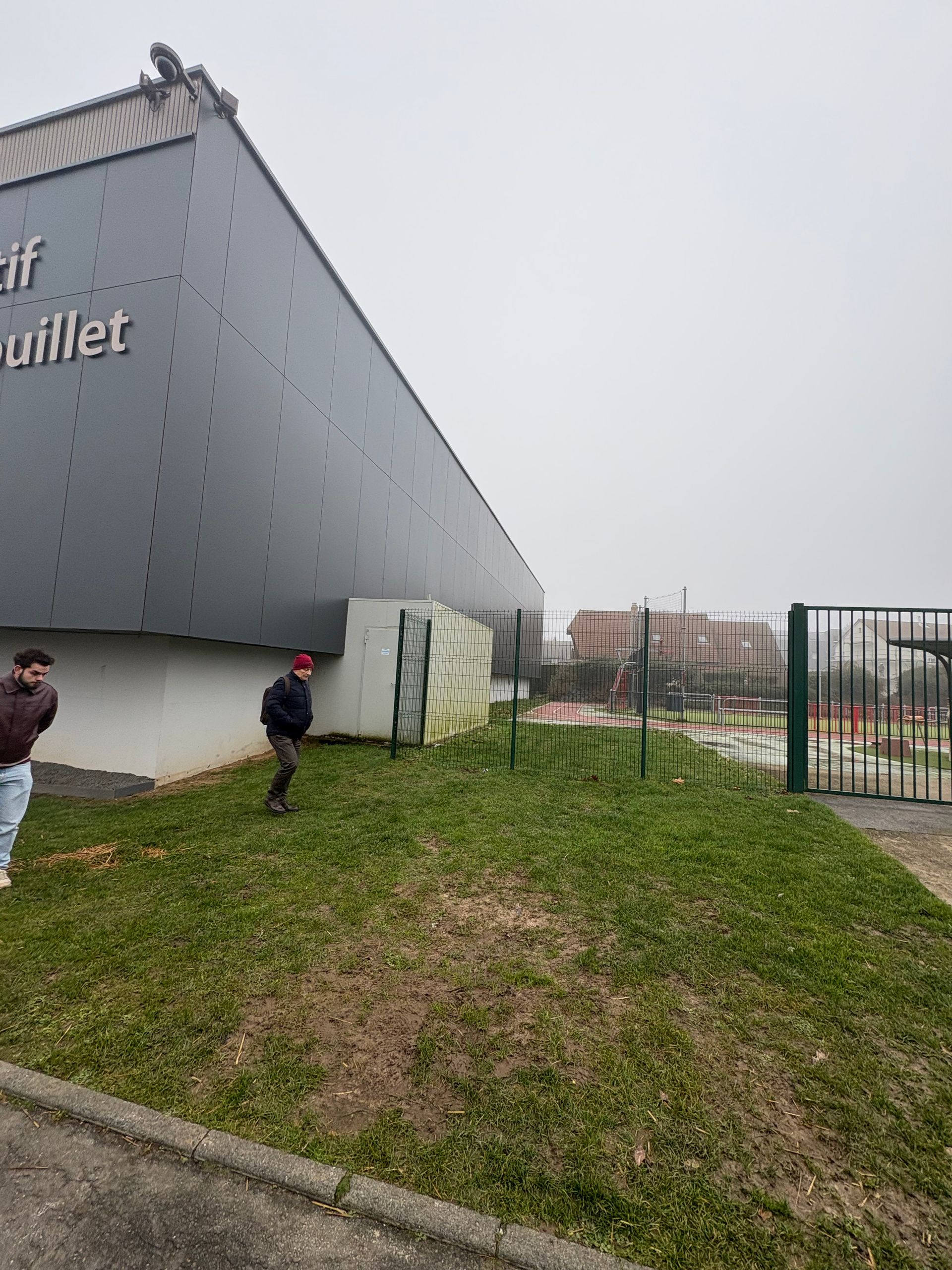 Deux personnes se tiennent debout sur une pelouse, à côté d'un grand bâtiment industriel gris et d'une clôture métallique, par une journée nuageuse.