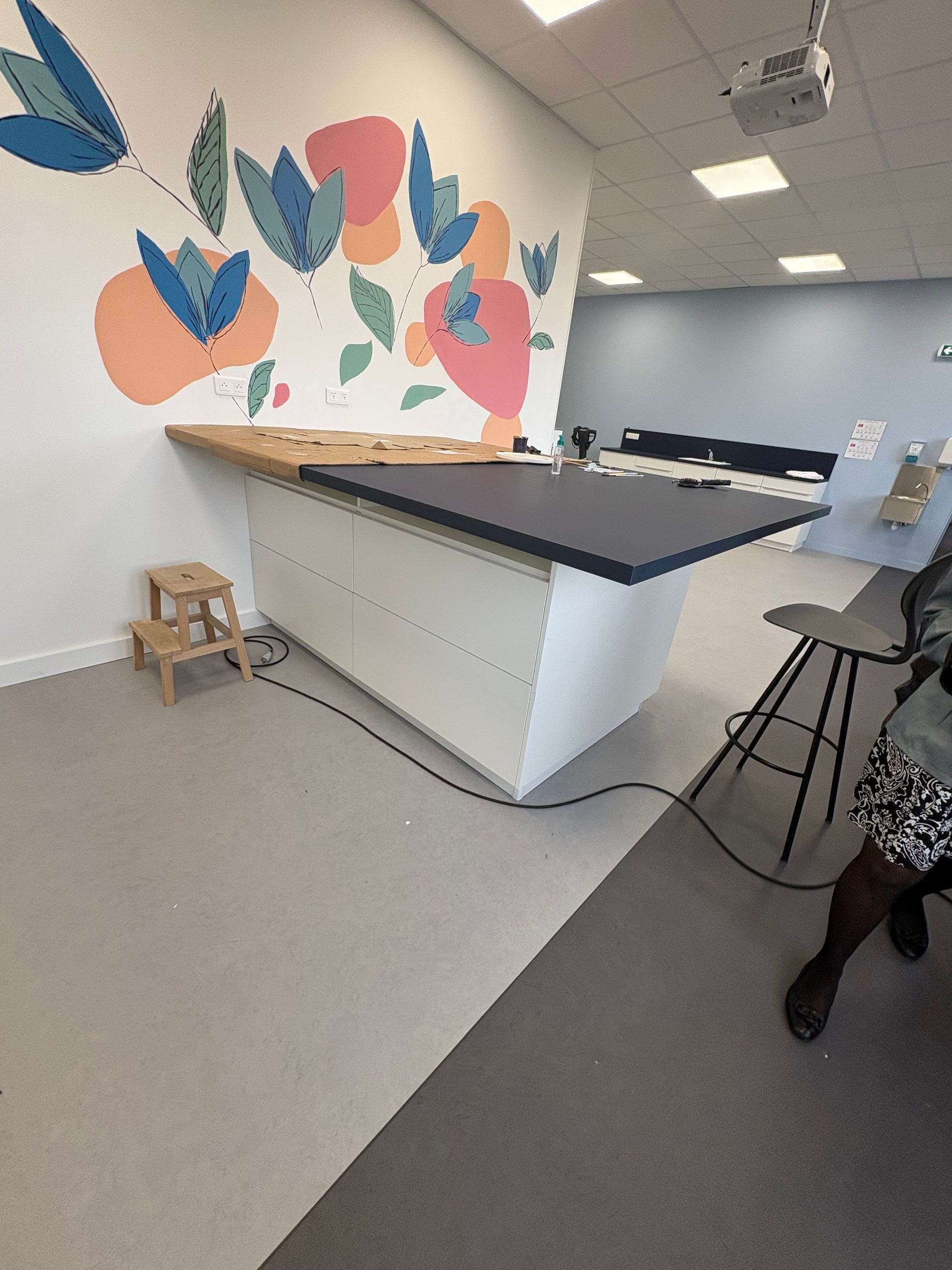 Dans un espace bureau, un îlot de cuisine blanc avec un plan de travail foncé est installé, avec une fresque florale au mur et un tabouret à proximité.
