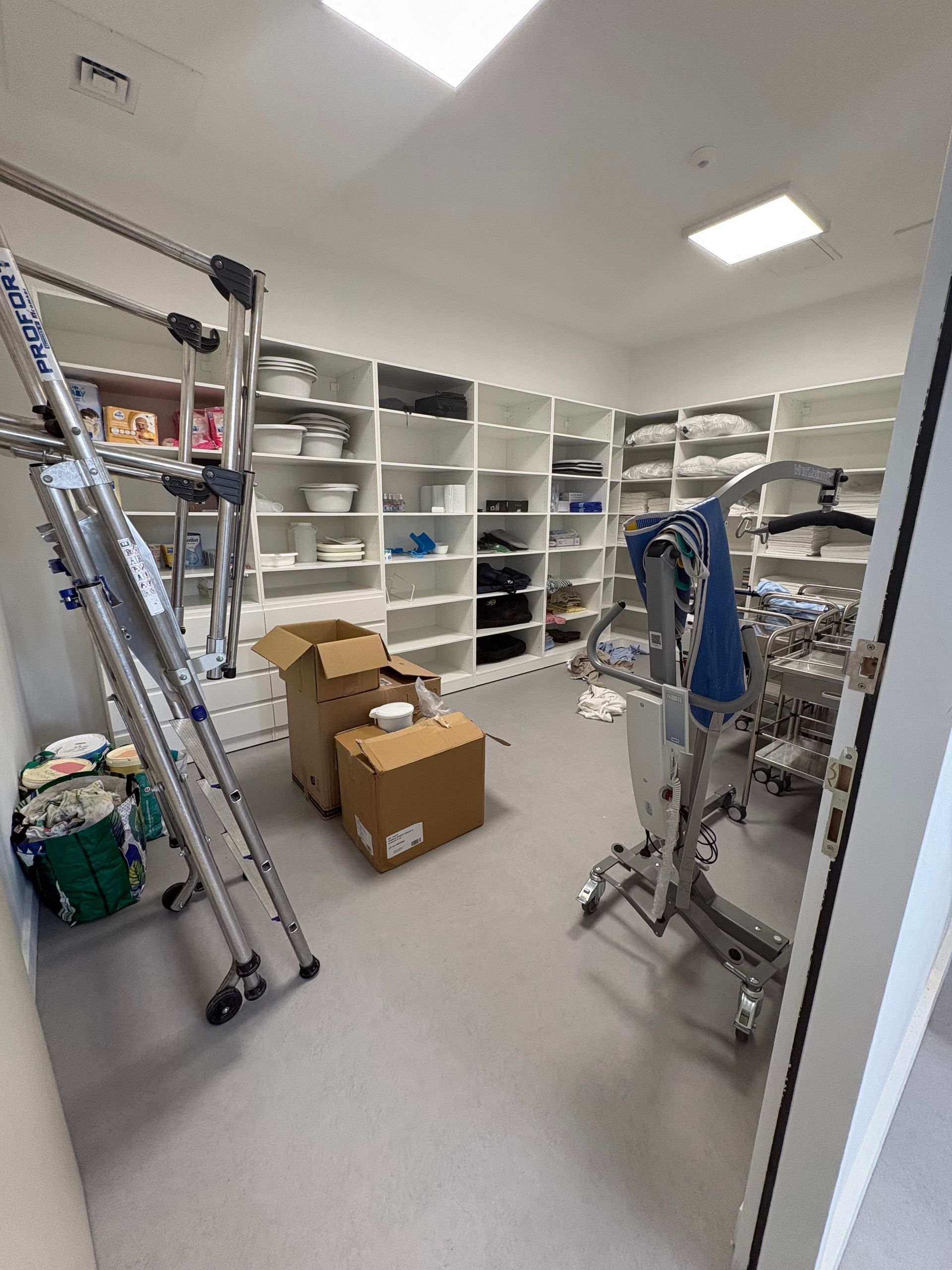 Un placard de rangement avec des étagères blanches contenant du matériel médical, deux cartons et deux lève-personnes.
