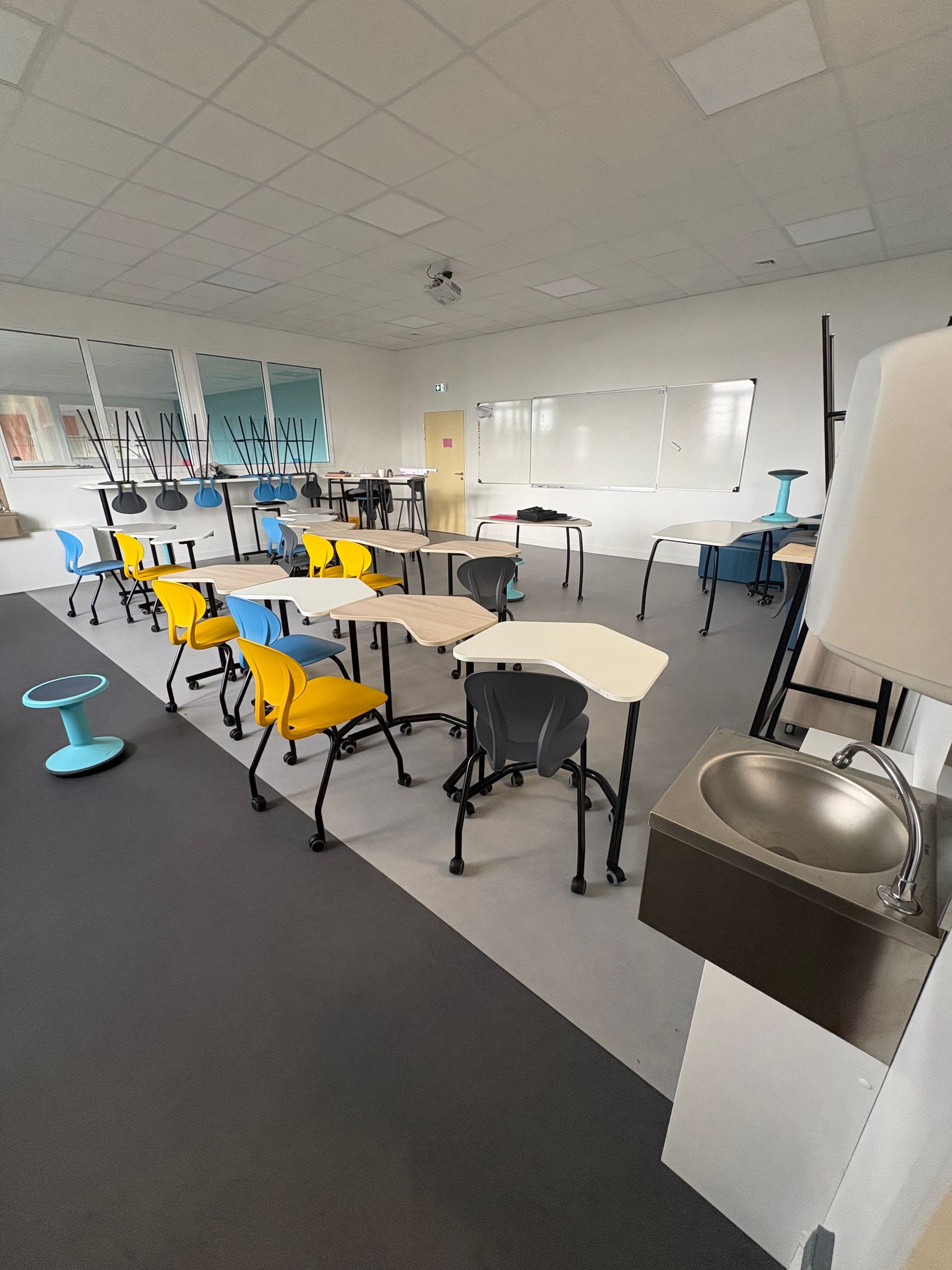 Une salle de classe avec des tables et des chaises colorées disposées de manière flexible, avec un lavabo au premier plan.