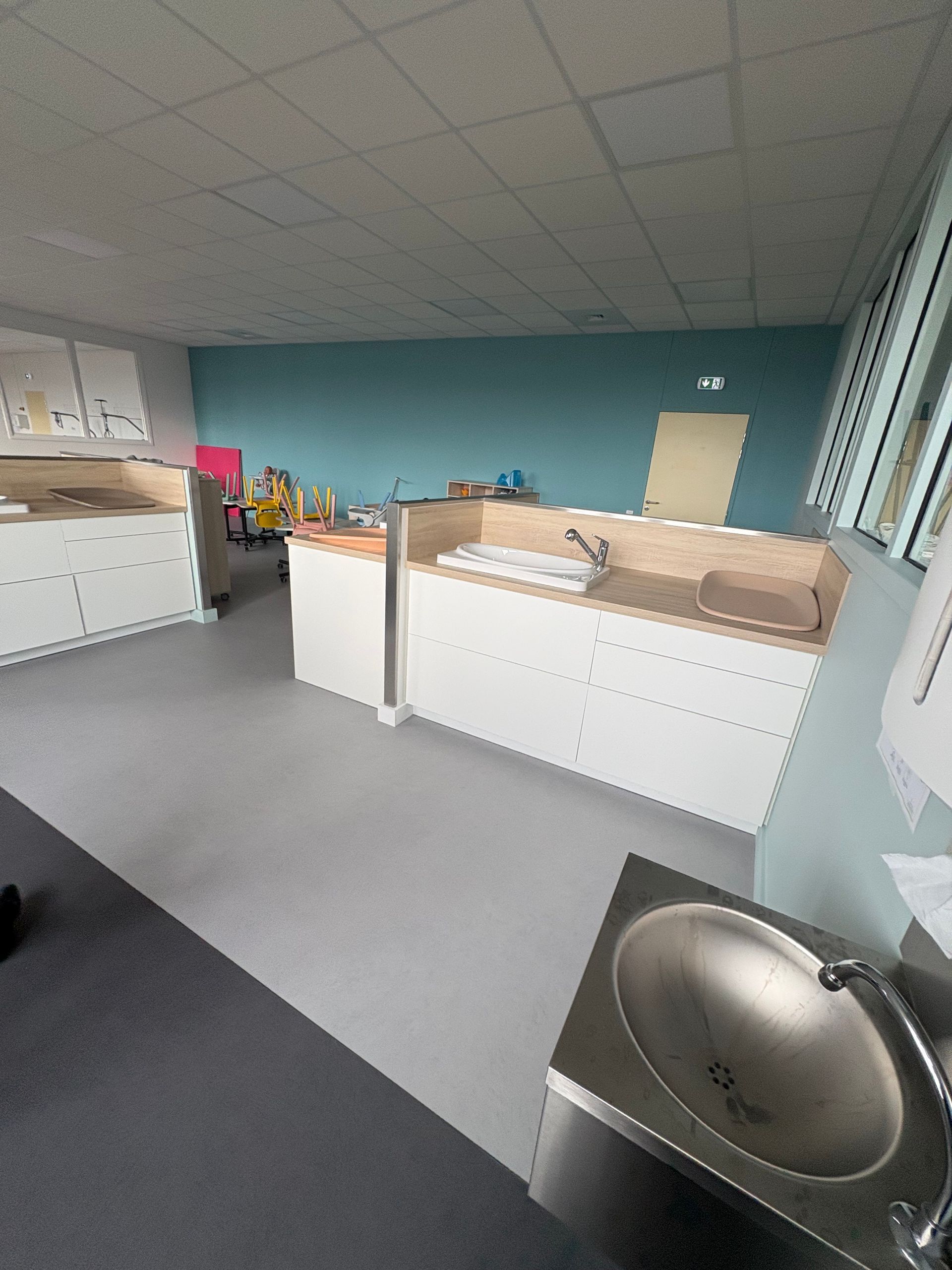 Une salle de garde d'enfants avec une table à langer blanche, un lavabo en métal, un sol gris et des murs aux accents turquoise.