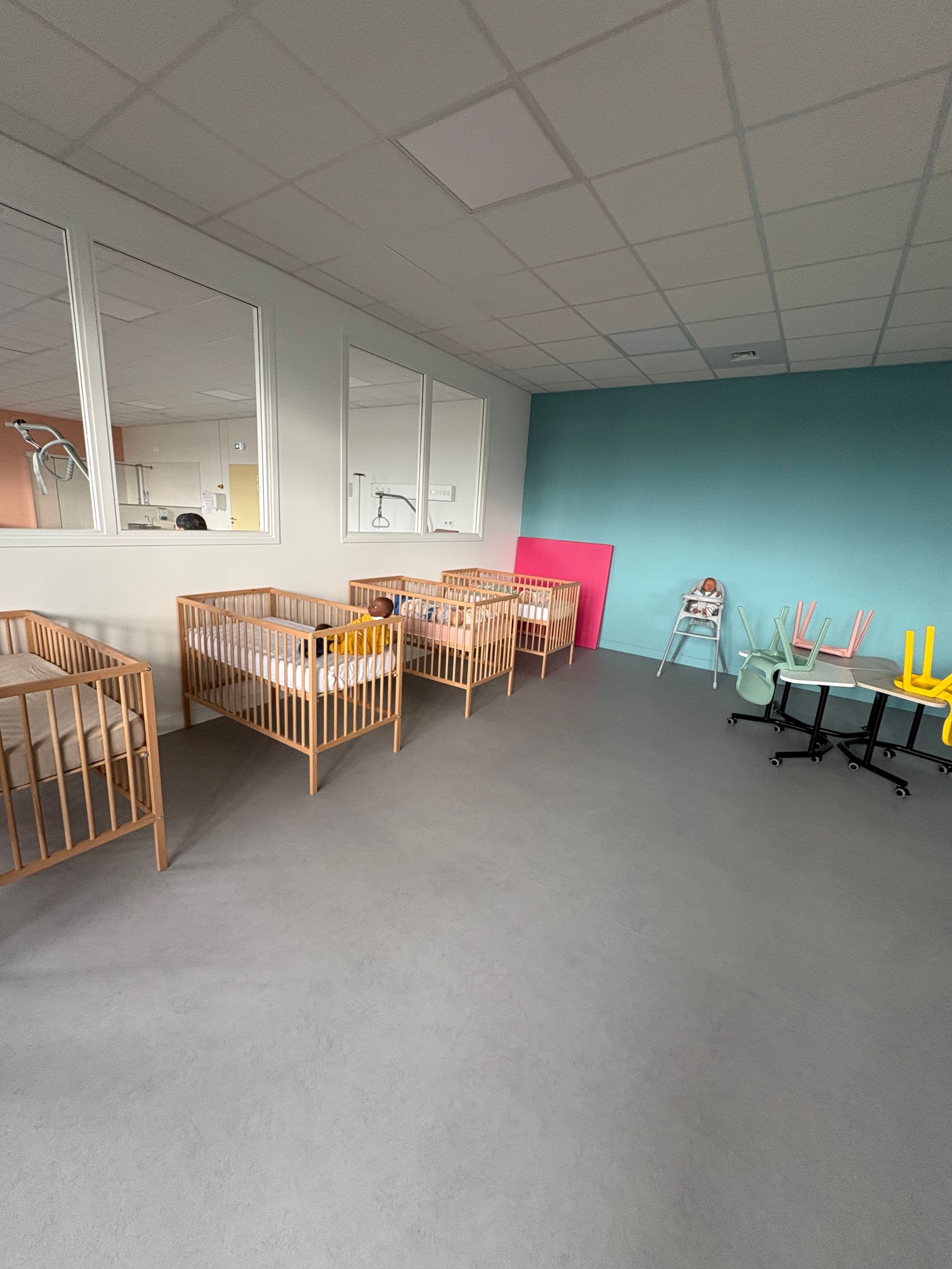 Une chambre d'enfant avec plusieurs berceaux en bois alignés, une chaise haute et des petites tables contre un mur turquoise.