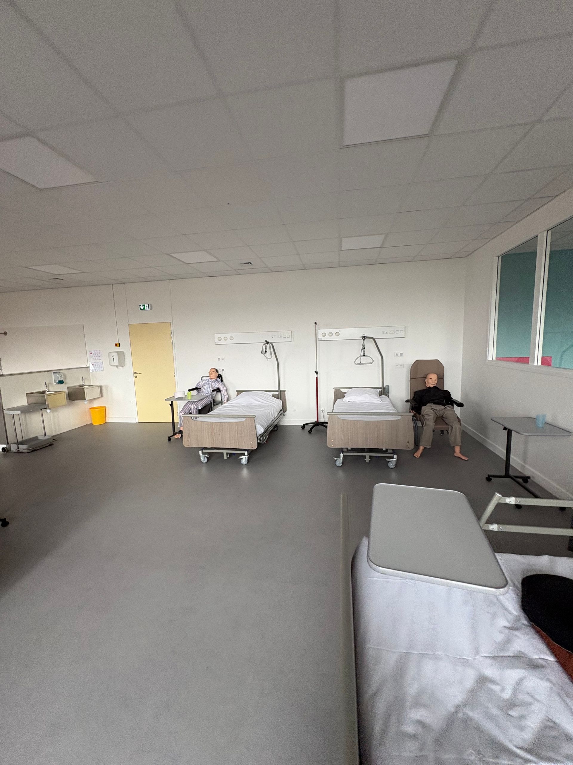Deux patients occupent une chambre d'hôpital avec deux lits et une chaise médicale, adossée à un mur blanc uni.