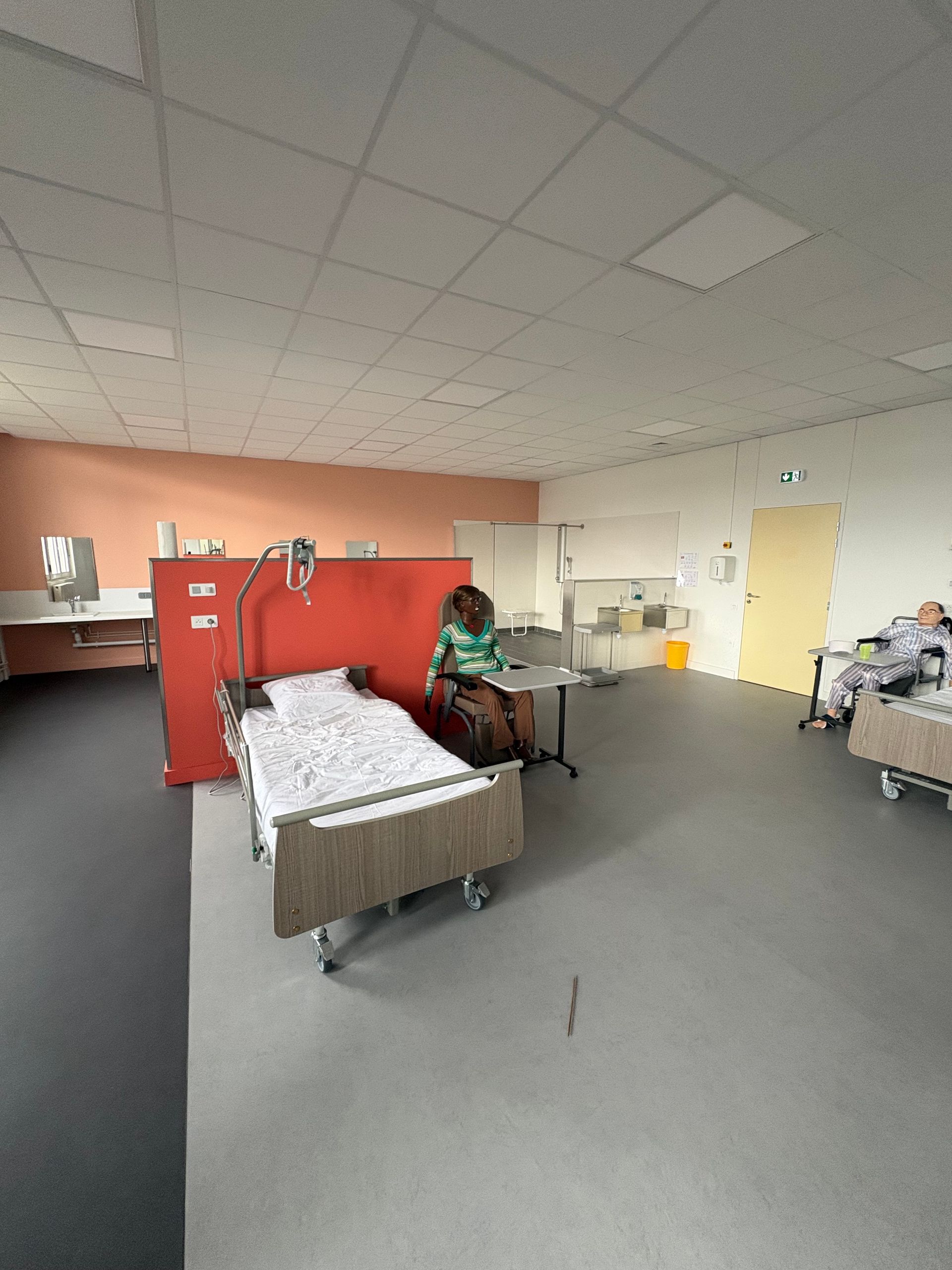 Plan large d'une chambre d'hôpital avec deux lits, une cloison rouge et une personne assise près d'une petite table.