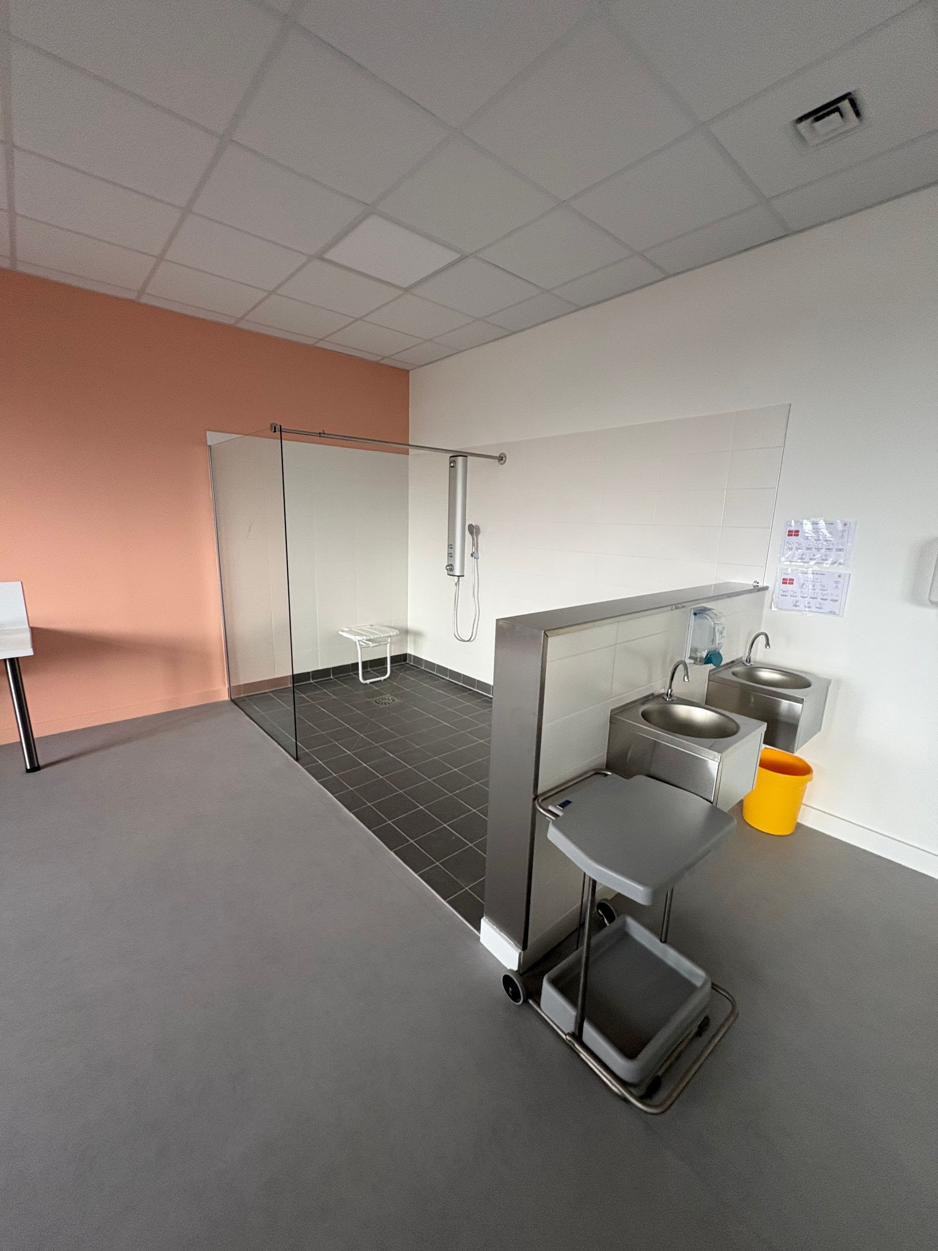 Des toilettes accessibles avec une cabine de douche carrelée, des barres d'appui, un lavabo en acier inoxydable et un sol gris.