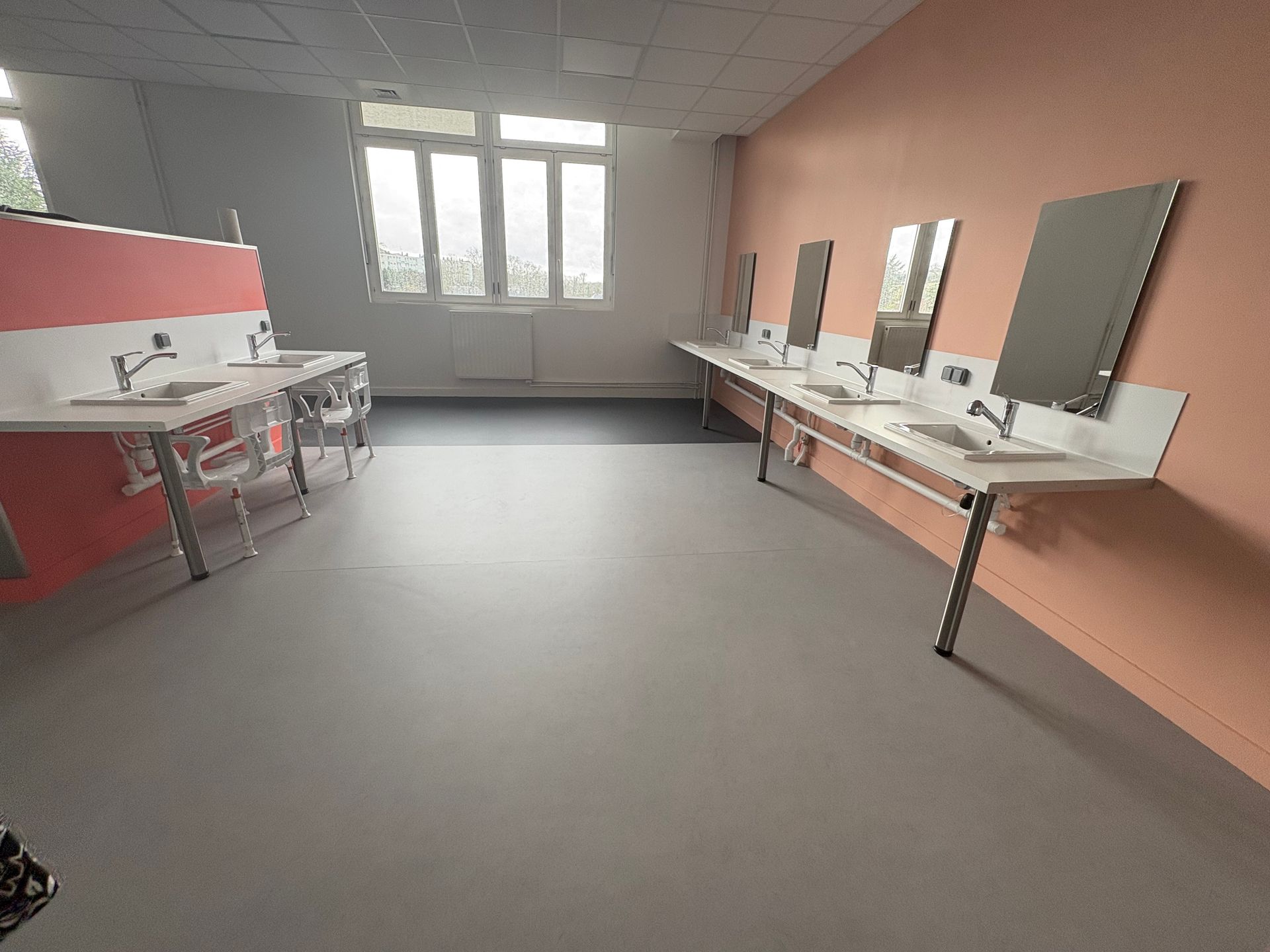 Une salle de bains lumineuse et vide, avec des rangées de lavabos blancs fixés au mur, des miroirs et des murs roses sur un sol gris.