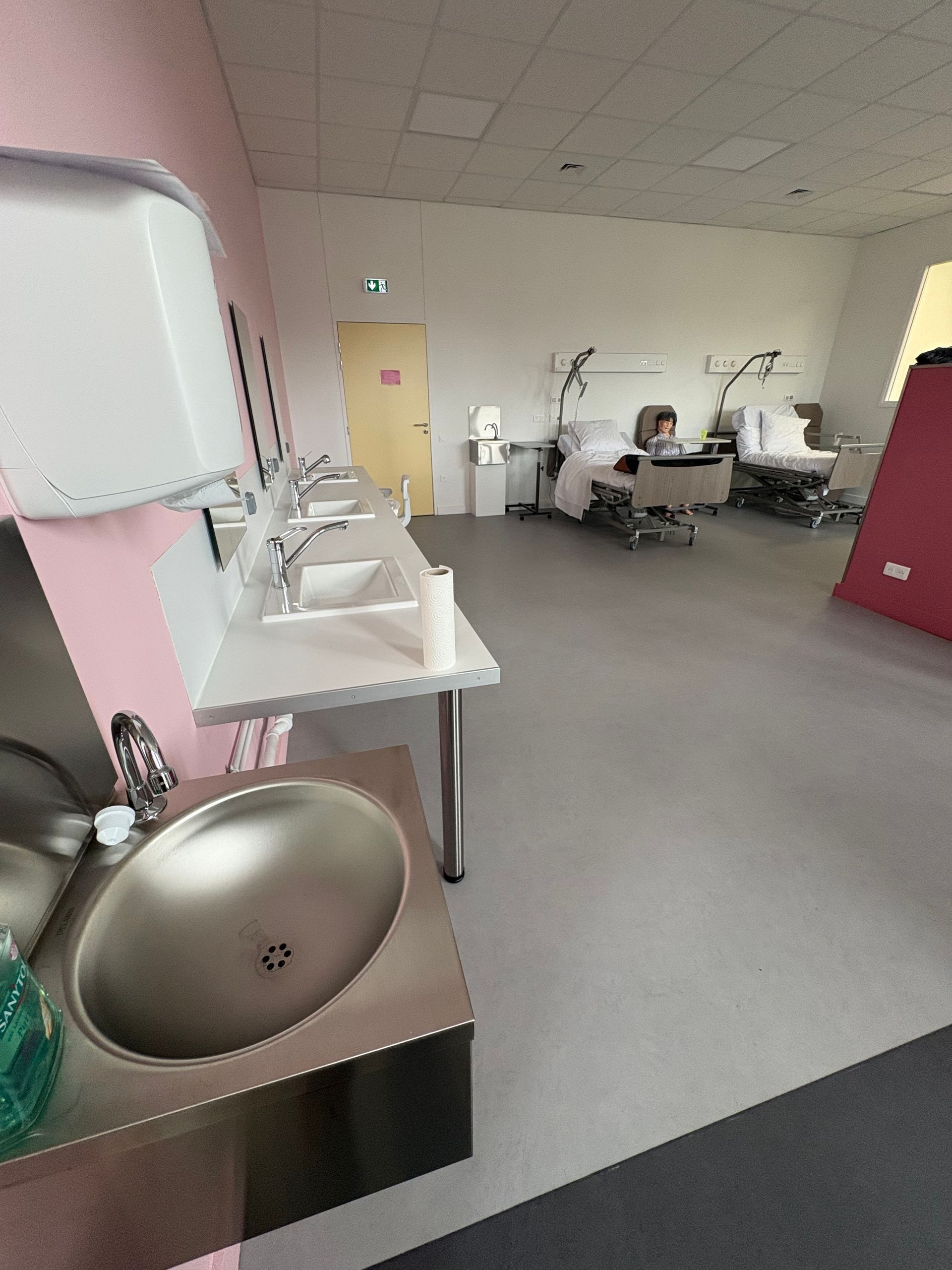 Une salle de soins avec un lavabo au premier plan, une rangée de lavabos muraux et deux lits médicaux à l'arrière-plan.