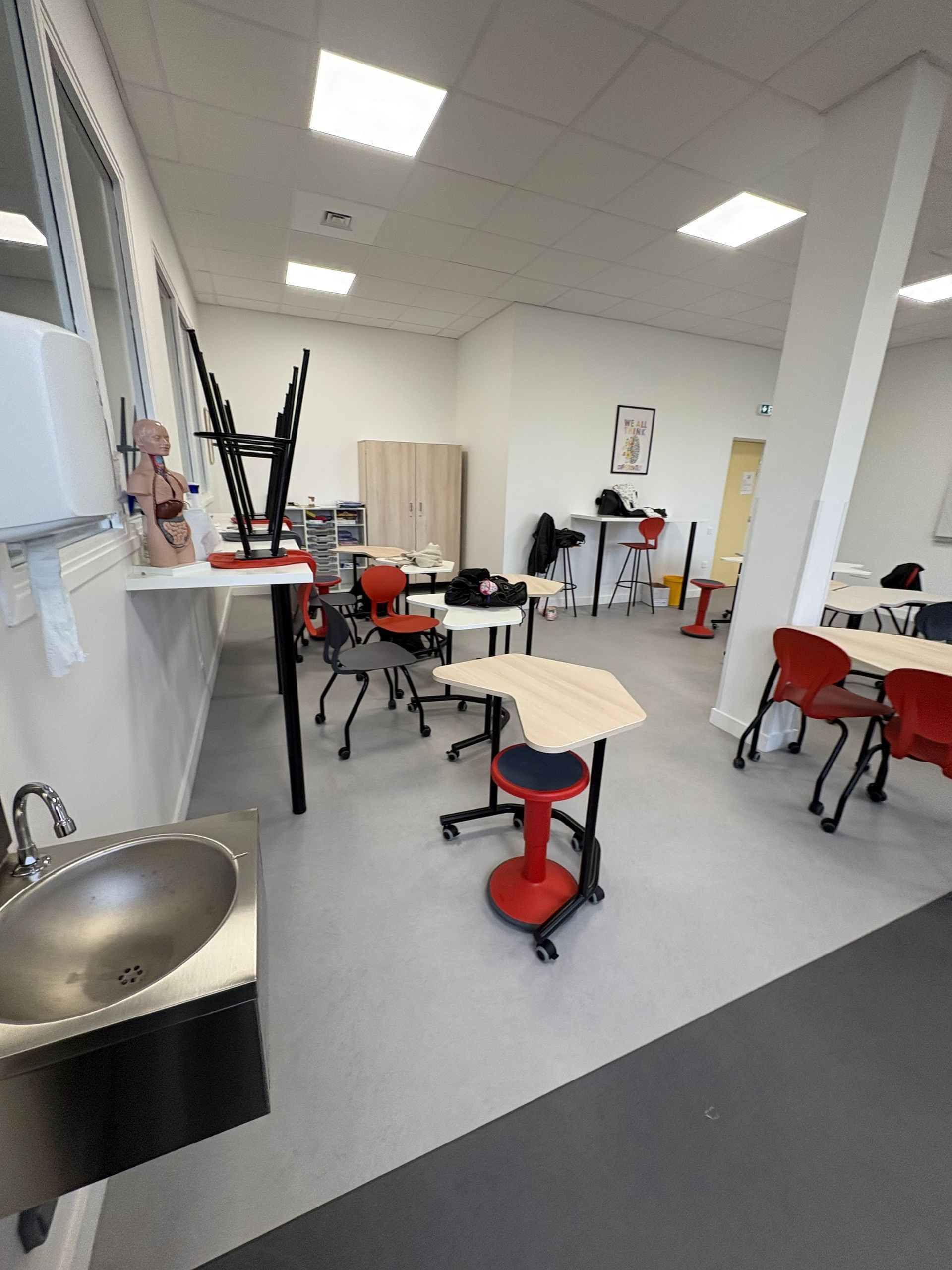 Une salle de classe avec des tables mobiles et des tabourets rouges, un lavabo et un distributeur d'essuie-mains en papier.