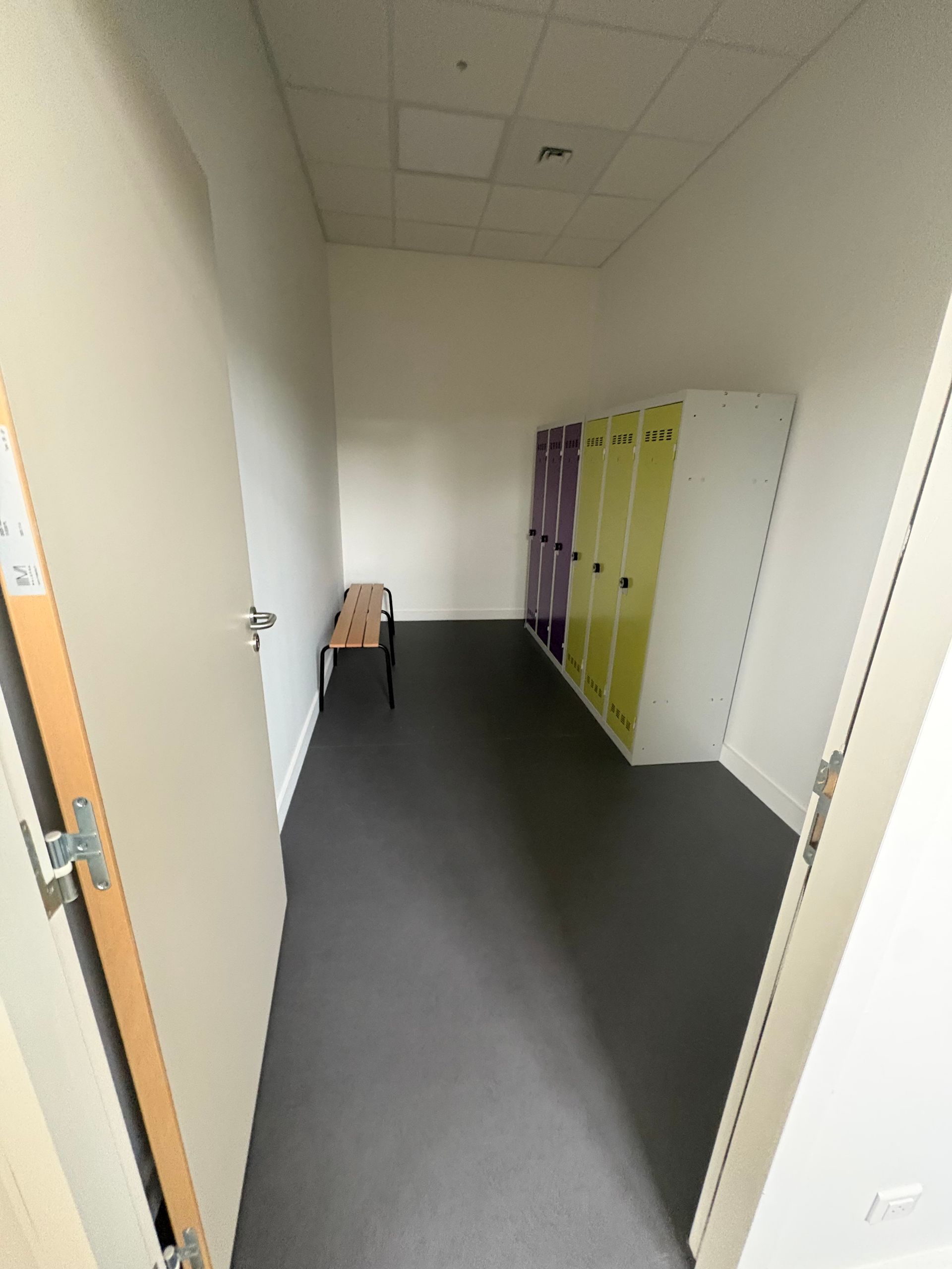 Un vestiaire étroit avec un banc contre le mur, une rangée de casiers verts et violets et un sol gris.