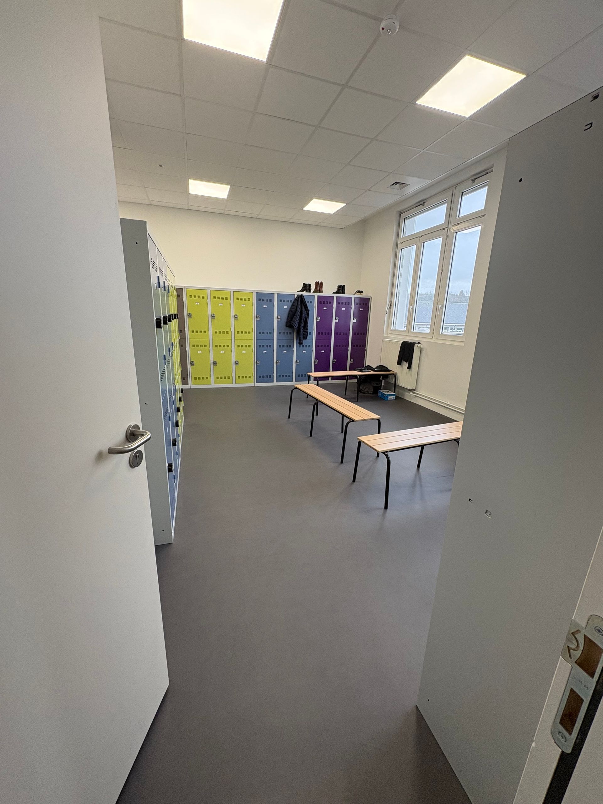 Un vestiaire avec des casiers colorés, deux bancs en bois au centre et une grande fenêtre sur un mur blanc.