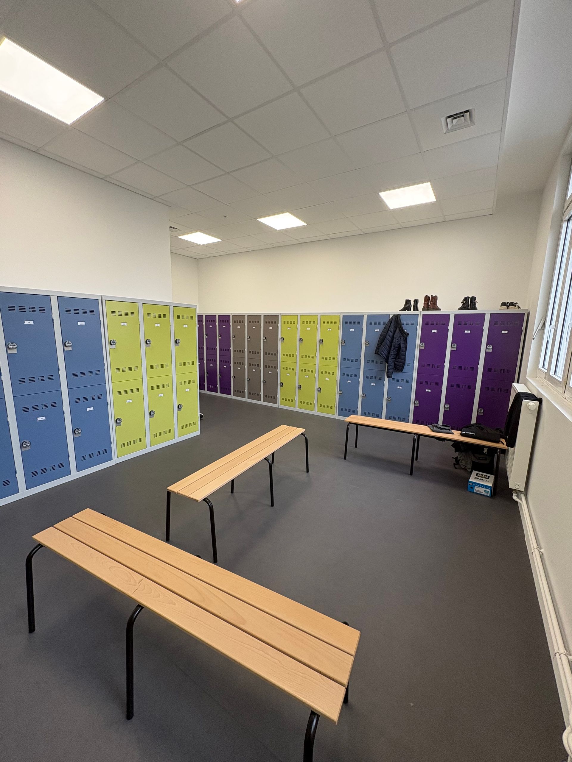Un vestiaire avec des rangées de casiers bleus, verts et violets, de la moquette grise et deux bancs en bois au centre.