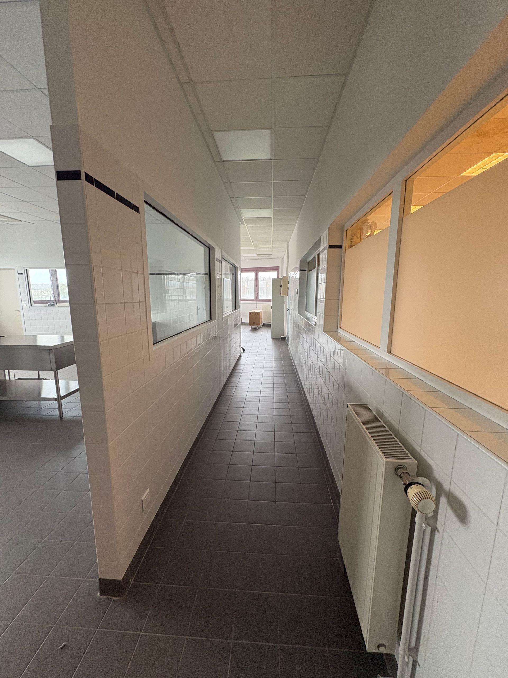 Un couloir étroit carrelé, avec un mur blanc à gauche et des fenêtres à stores orange à droite, menant à une porte.