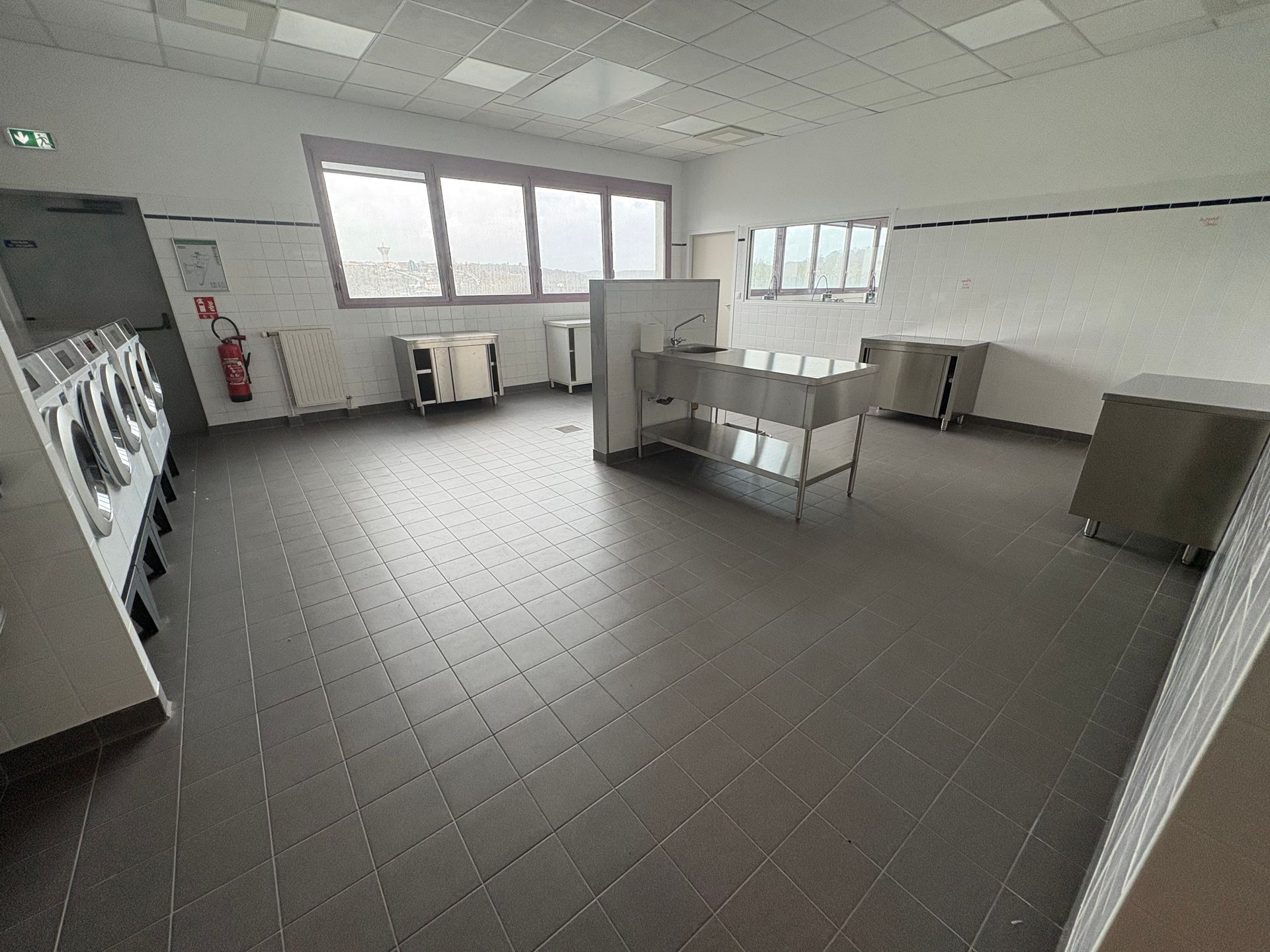 Une cuisine commerciale stérile et vide, avec un sol carrelé gris, des postes de travail en acier inoxydable et des murs blancs.