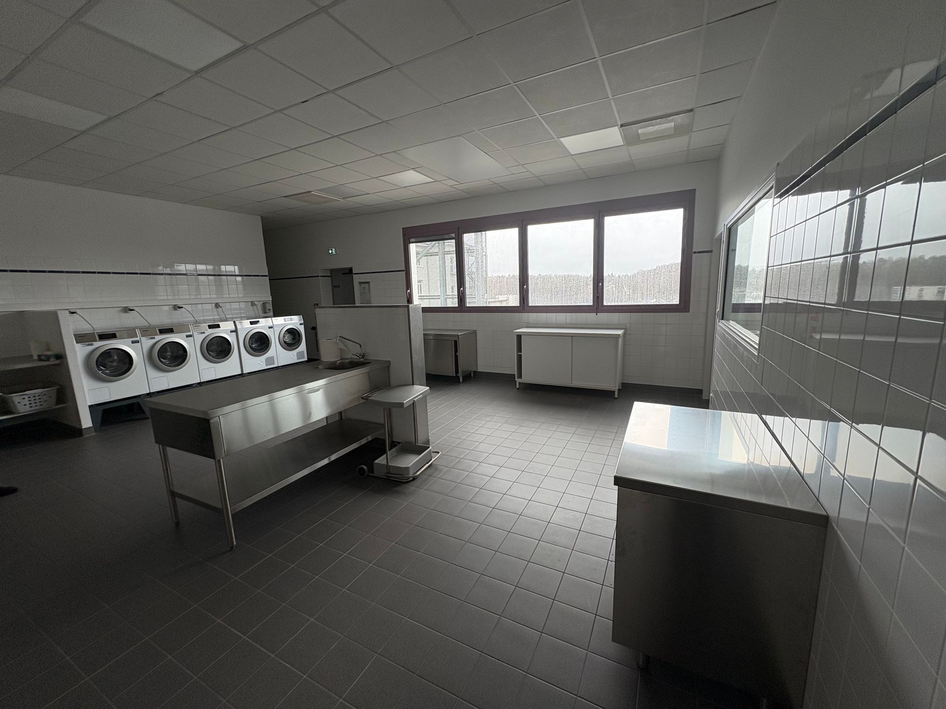 Une buanderie lumineuse et carrelée, avec une rangée de machines à laver, une table pliante en métal et de grandes fenêtres.