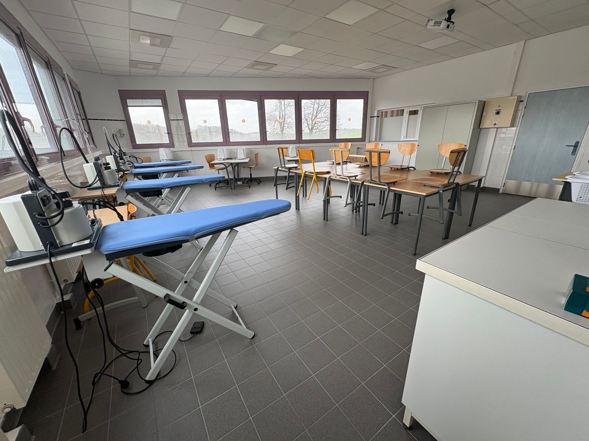 Une buanderie ou une salle de classe textile comprenant plusieurs tables à repasser recouvertes de housses bleues et des rangées de chaises et de tables.