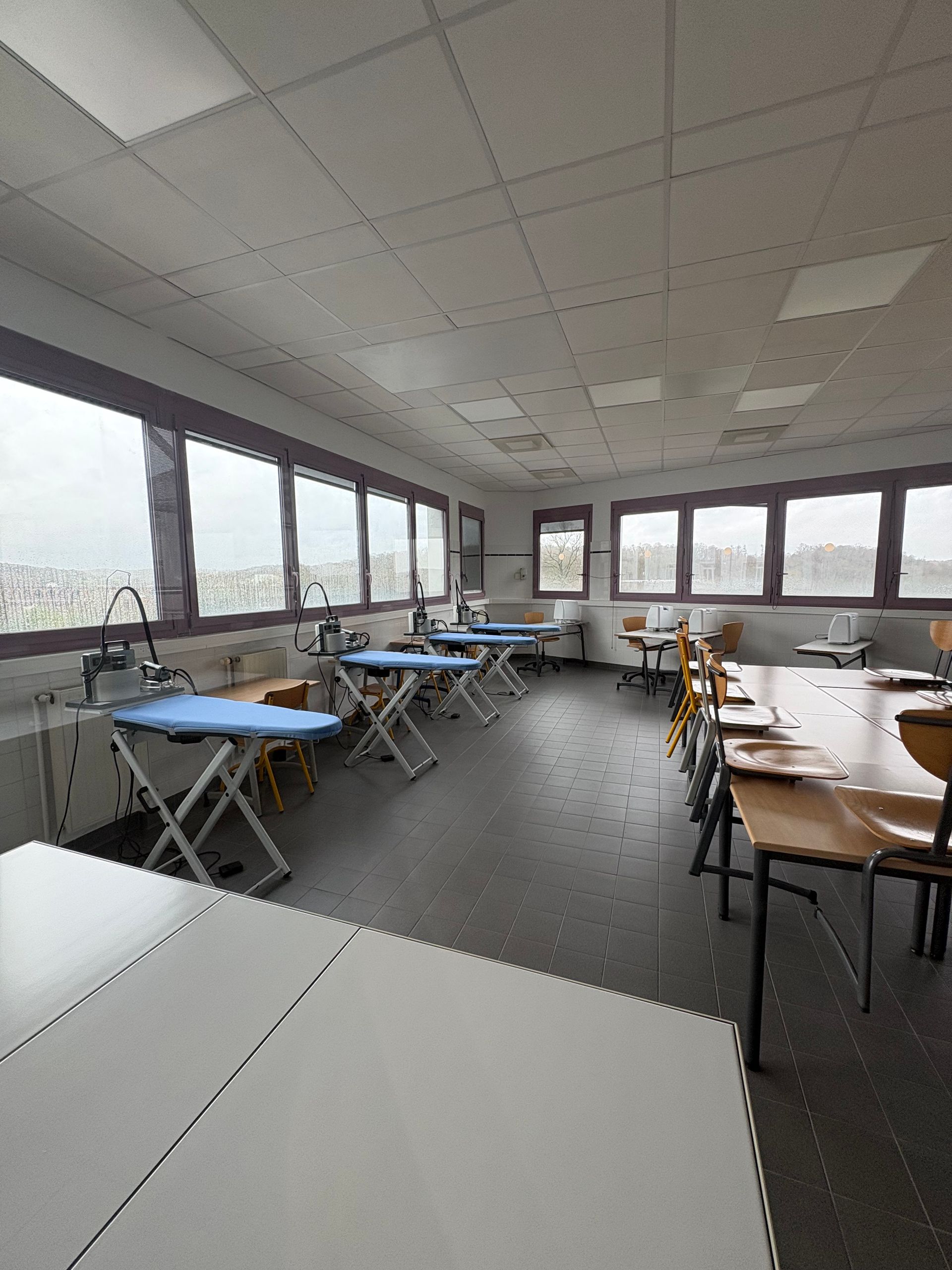 Une salle de classe lumineuse avec plusieurs planches à repasser alignées près de grandes fenêtres et une grande table sur la droite.