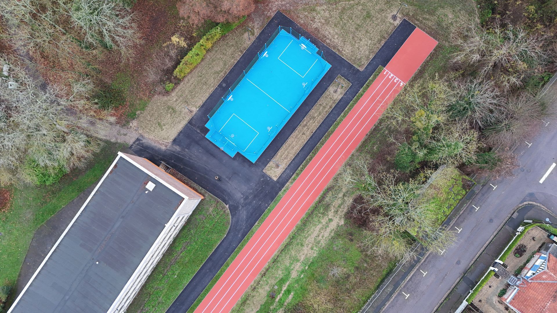 Vue aérienne d'une cour d'école montrant un terrain de sport rectangulaire bleu, une piste d'athlétisme rouge et le toit d'un bâtiment.