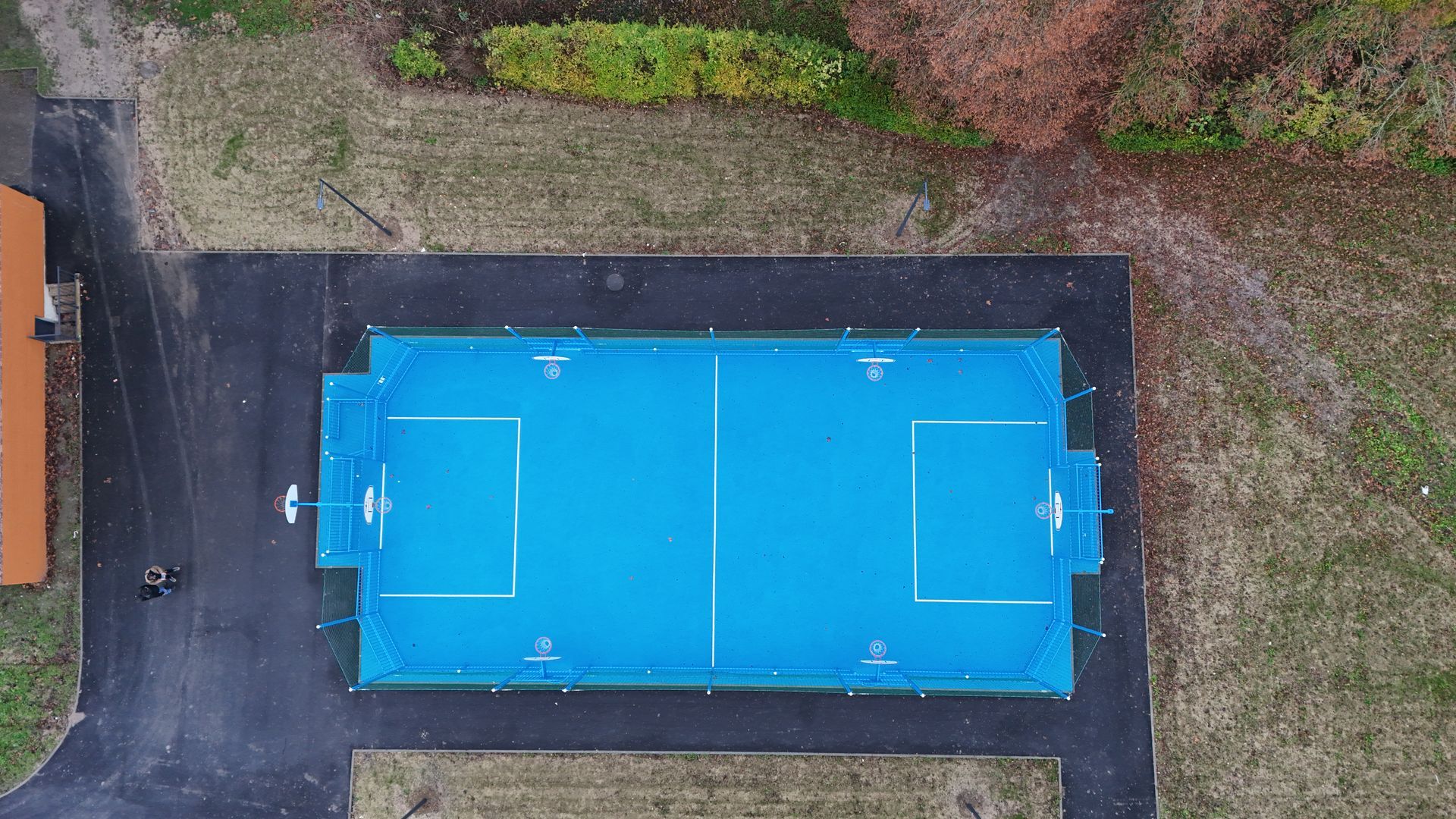 Vue aérienne d'un terrain de football à effectif réduit, d'un bleu vif, sur une surface pavée sombre et entouré d'herbe.