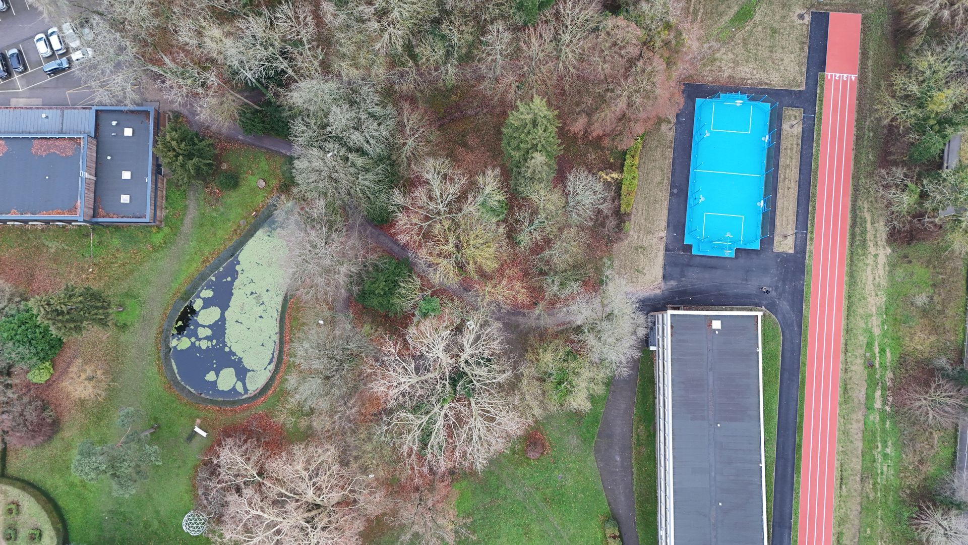 Vue aérienne d'un campus avec un étang, un bâtiment rectangulaire, une structure recouverte de bleu et une piste d'athlétisme rouge.