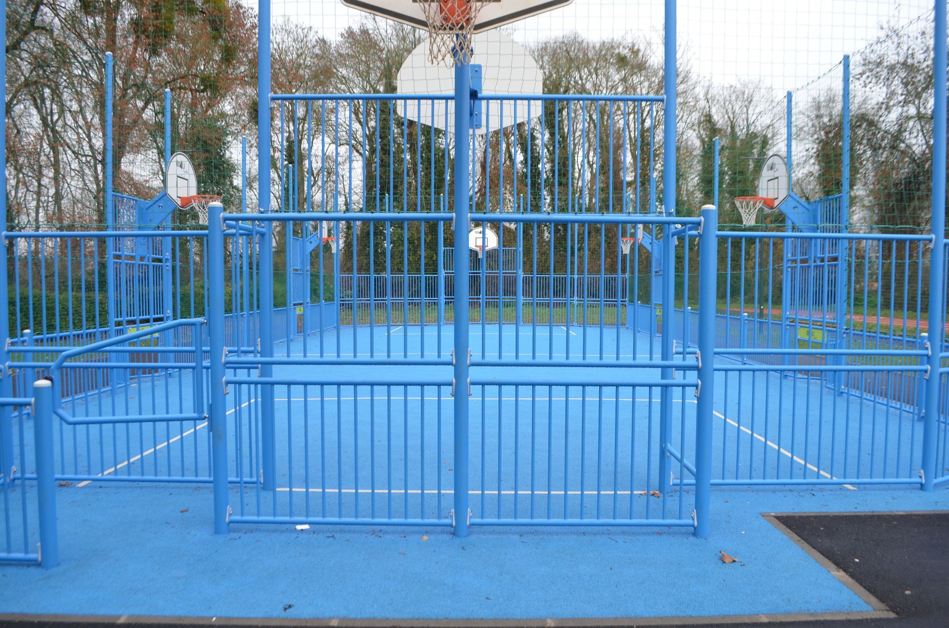 Un terrain de basket-ball bleu vif, clôturé, avec deux paniers, entouré d'arbres.