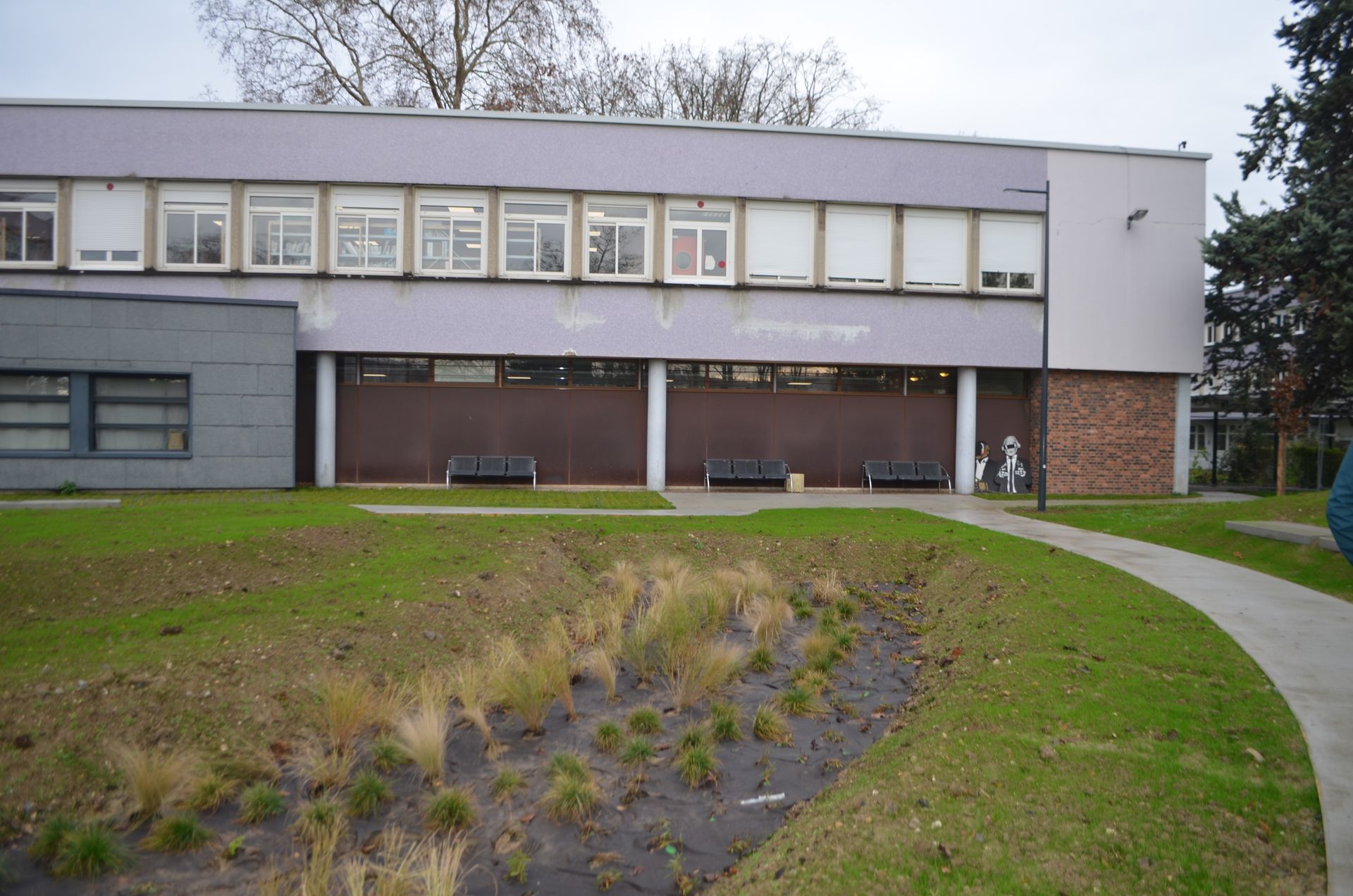 Un bâtiment de deux étages avec un revêtement extérieur violet clair et de nombreuses fenêtres, donnant sur une pelouse agrémentée d'un parterre de jardin paysager.