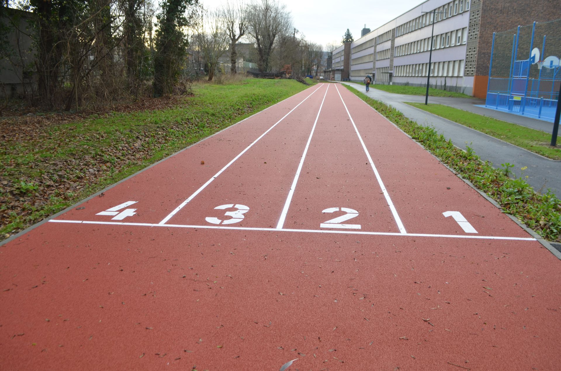 Une piste d'athlétisme rouge à quatre couloirs, numérotée de blanc, mène à un bâtiment scolaire situé dans un parc extérieur.