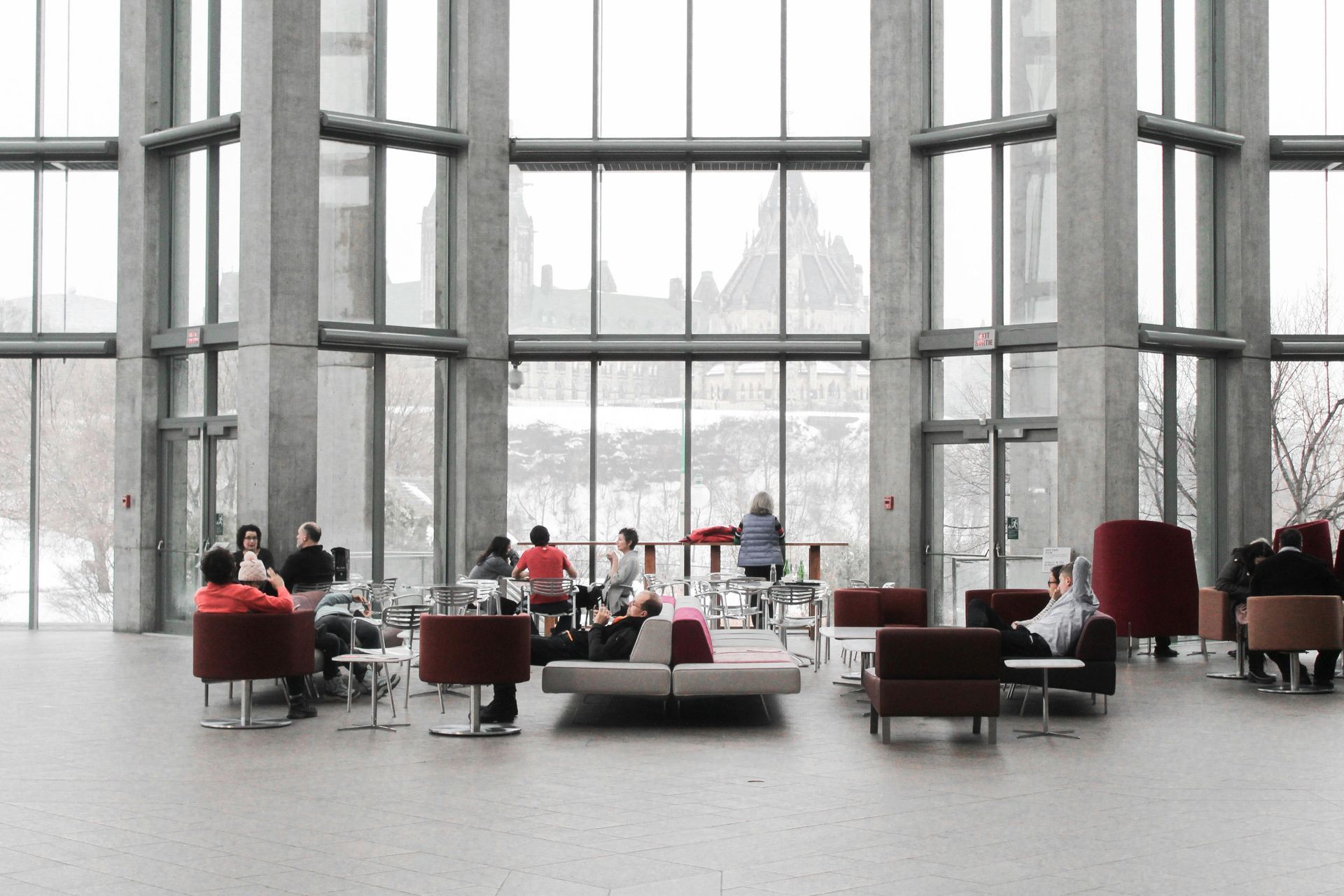 Des personnes sont assises dans un salon moderne aux parois de verre et aux piliers en béton, avec vue sur les édifices du Parlement canadien.