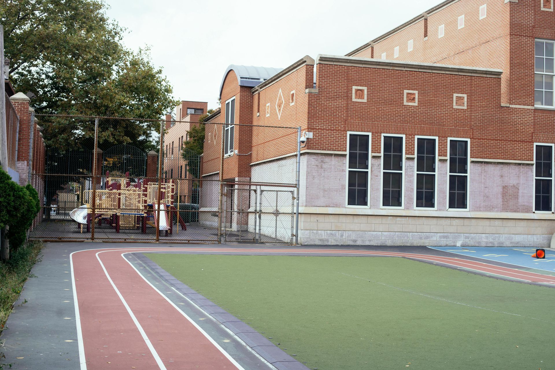 Un bâtiment scolaire en briques borde une cour de récréation clôturée, avec un terrain en gazon synthétique vert et une piste d'athlétisme rouge.