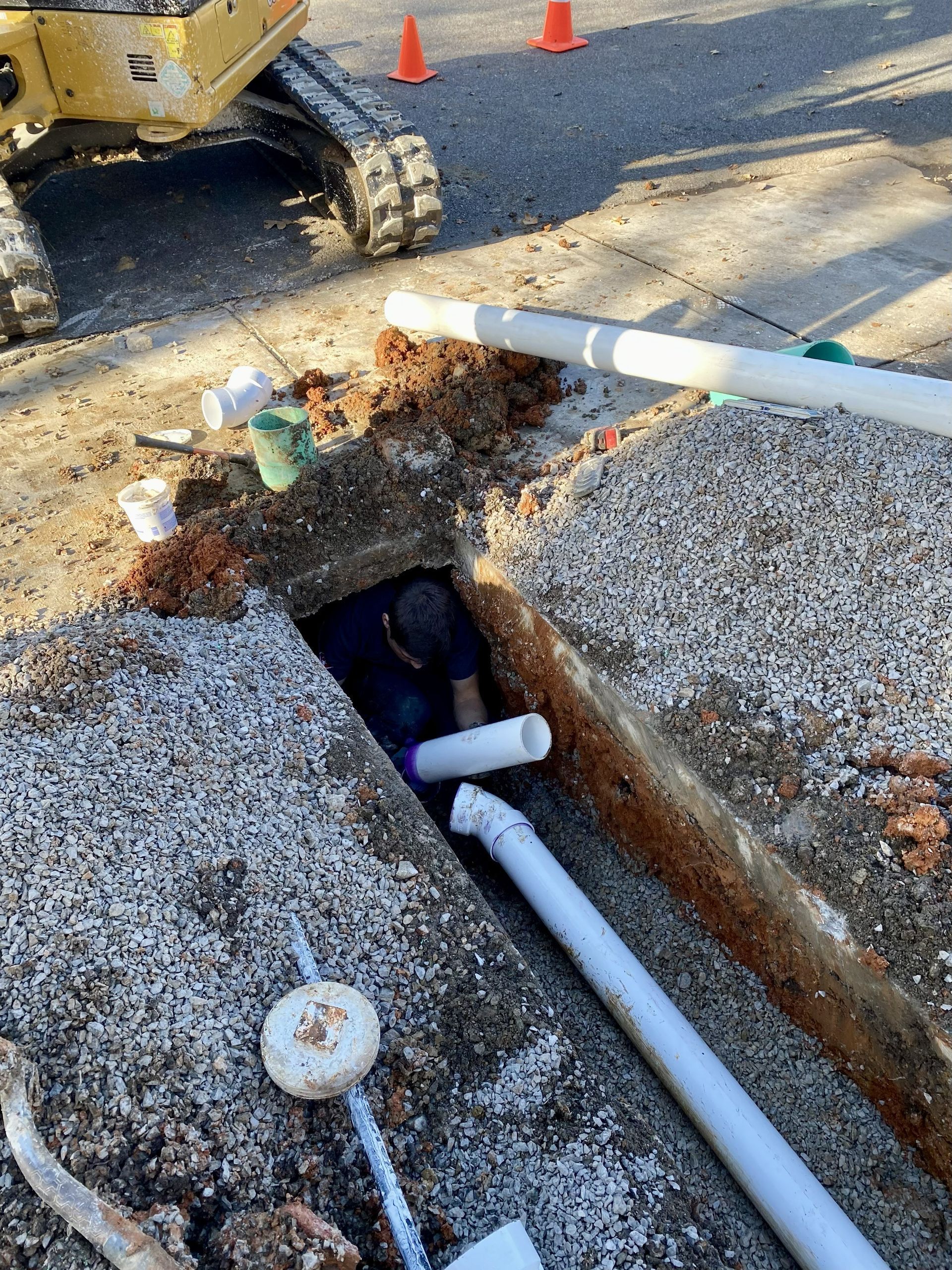 Construction worker in a trench repairing a pipe, with an excavator and orange cones nearby.