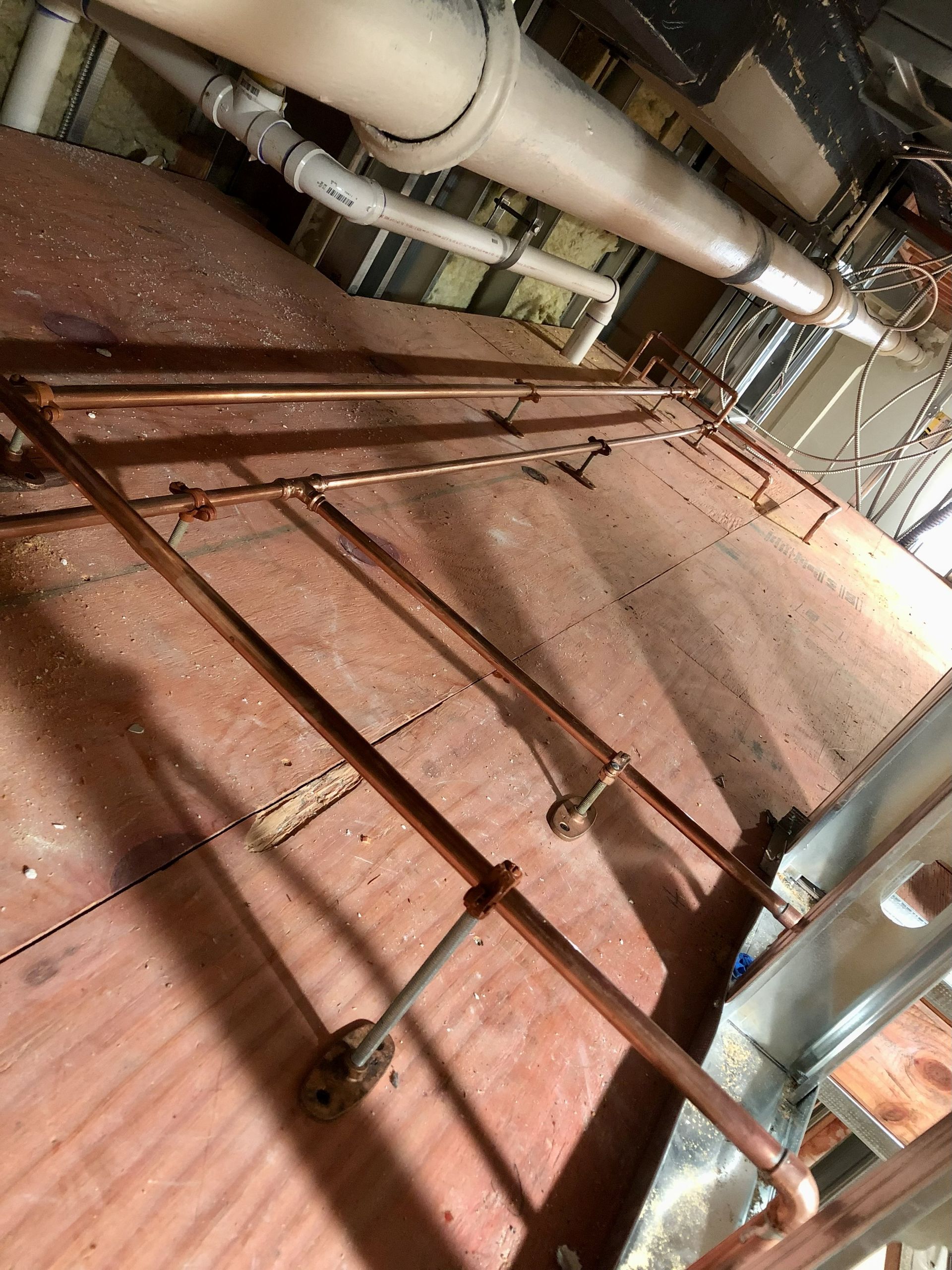 Copper pipes installed on a wooden floor in a building's interior. Other pipes and ceiling visible.