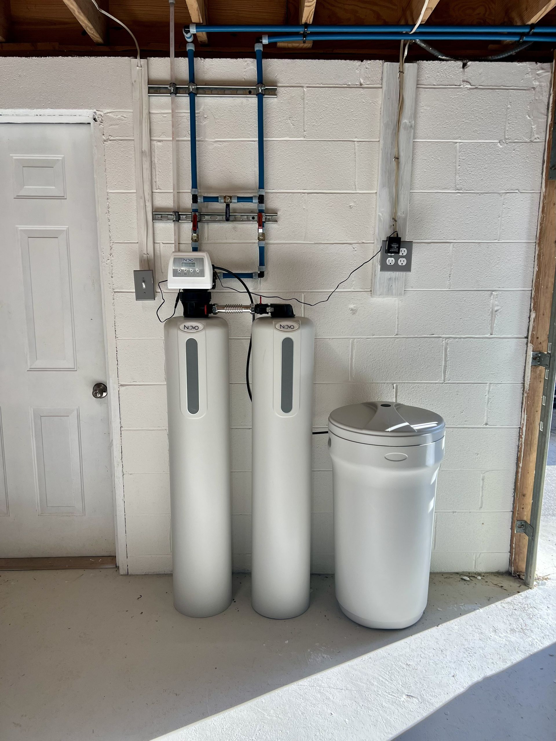 Water filtration system with three white tanks against a cinder block wall. Blue pipes are above.