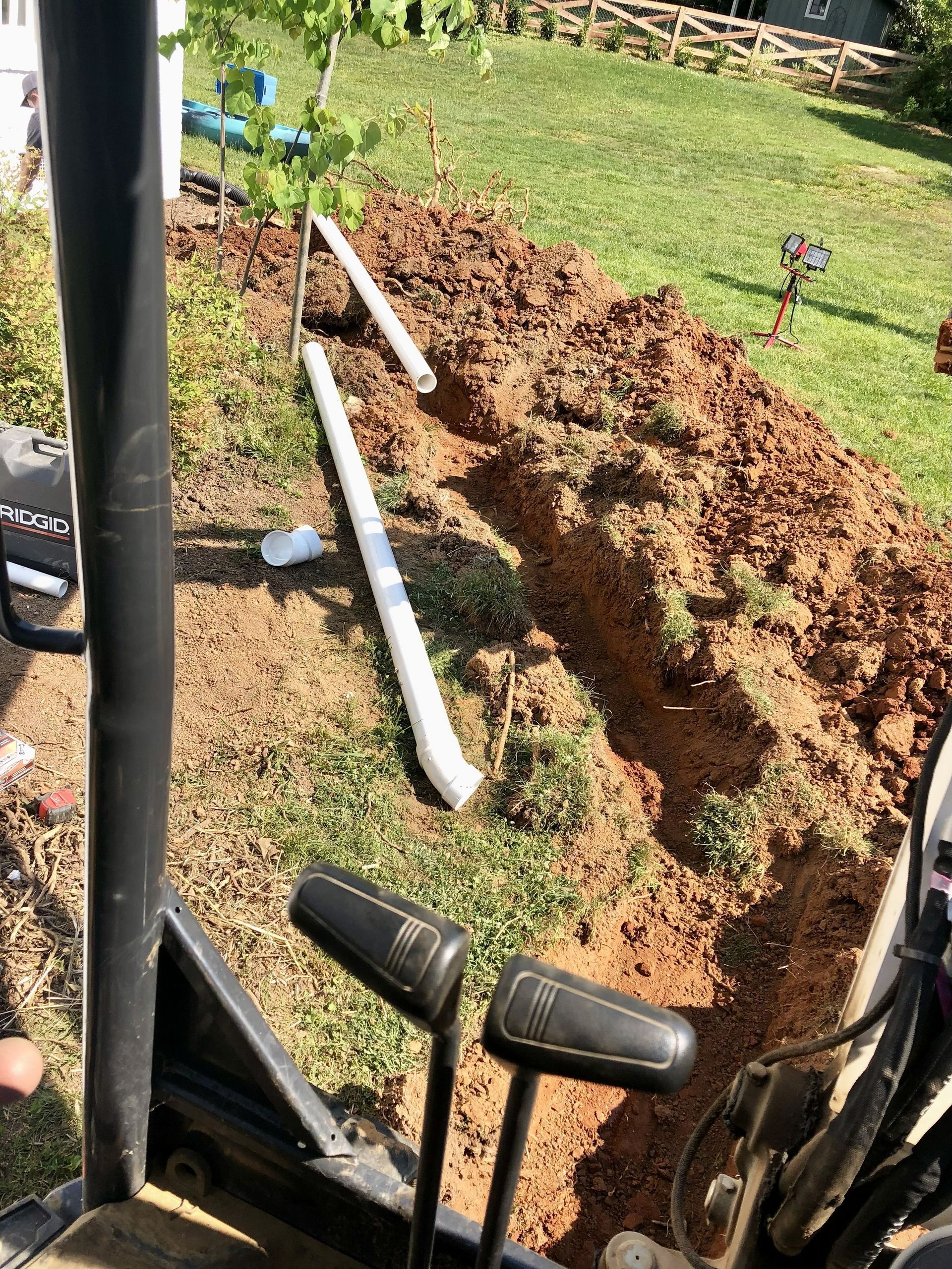 A trench in the brown earth with white pipes, seen from the controls of a machine outdoors.
