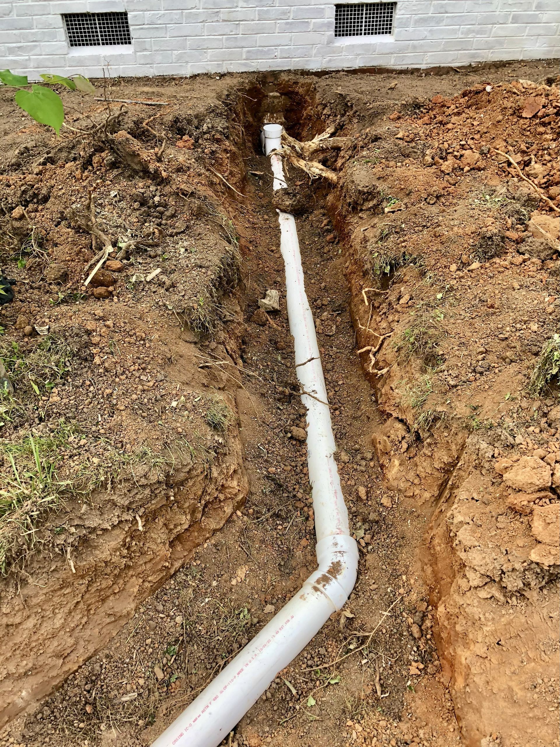 White PVC pipe in a trench beside a brick foundation. Brown dirt and green grass surround the trench.