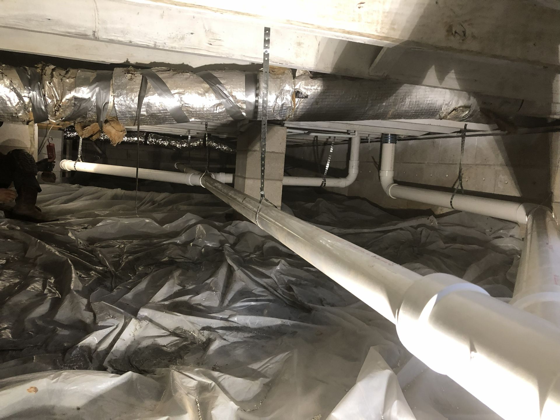 White pipes in a crawlspace under a structure with insulation, concrete, and plastic sheeting.