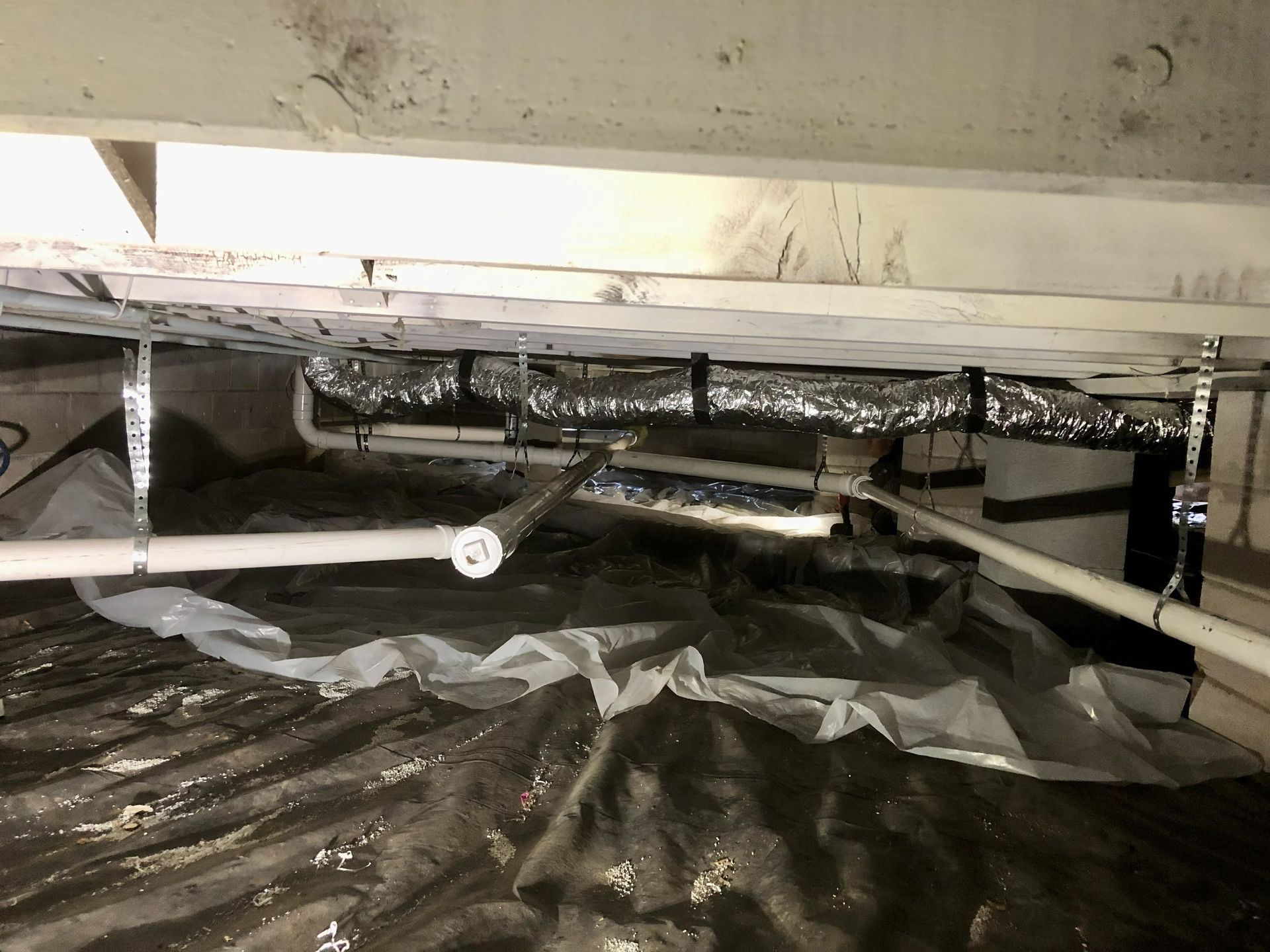 Crawl space with insulation, pipes, and ductwork beneath a house, covered with a black plastic vapor barrier.