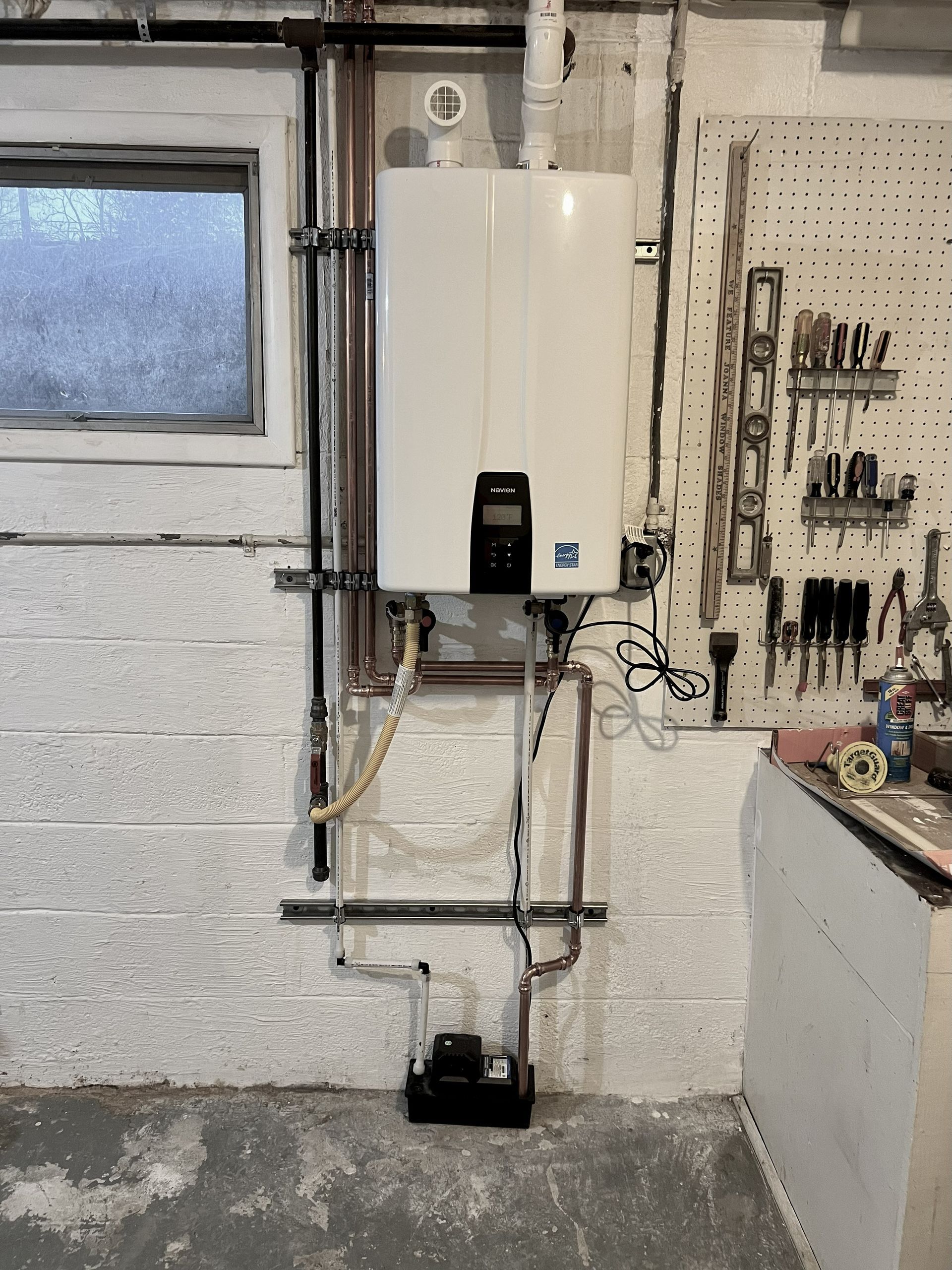 White tankless water heater on a basement wall with pipes and a pegboard of tools.