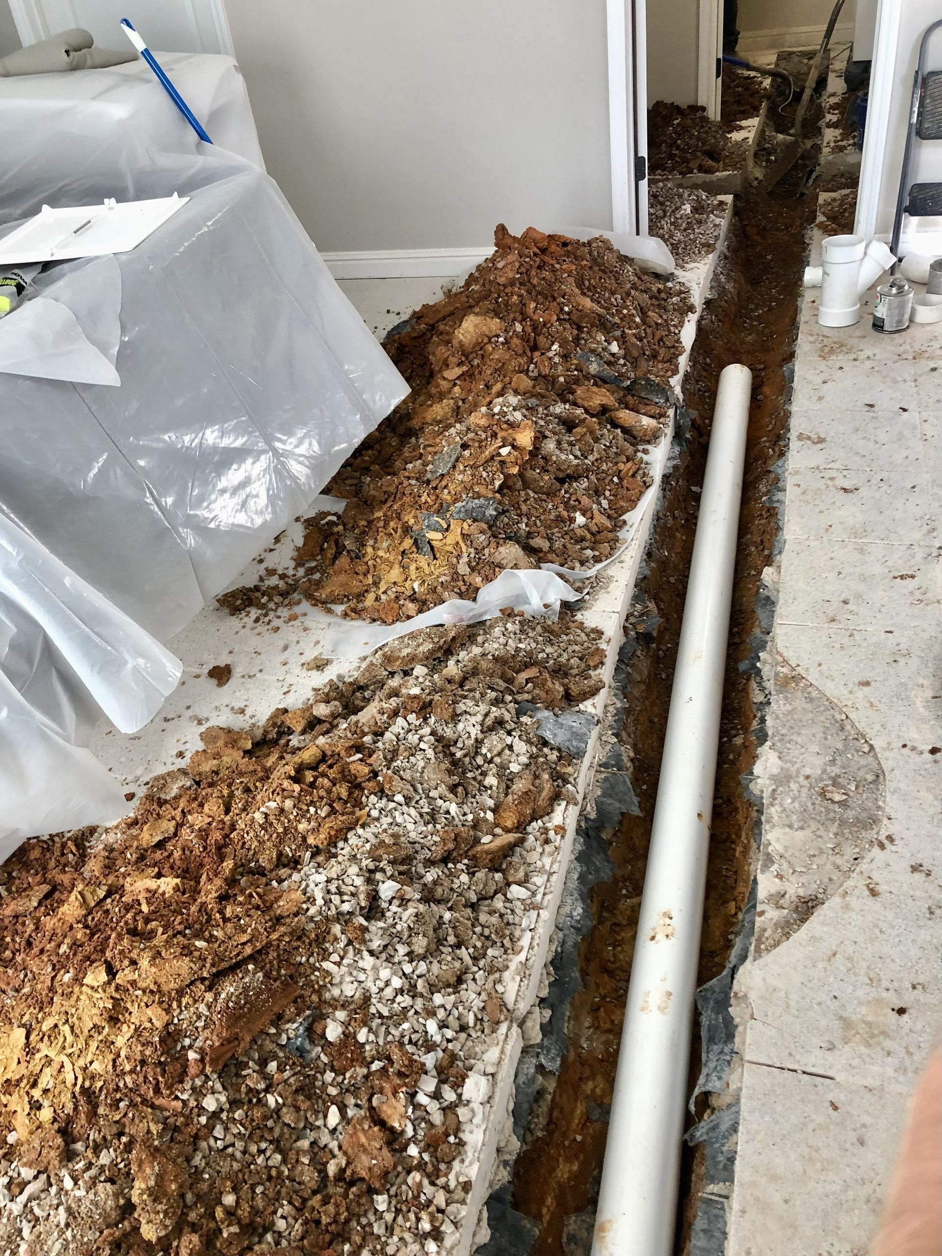 Trench dug in a concrete floor, with a white pipe laid inside. Brown soil piled alongside the trench.