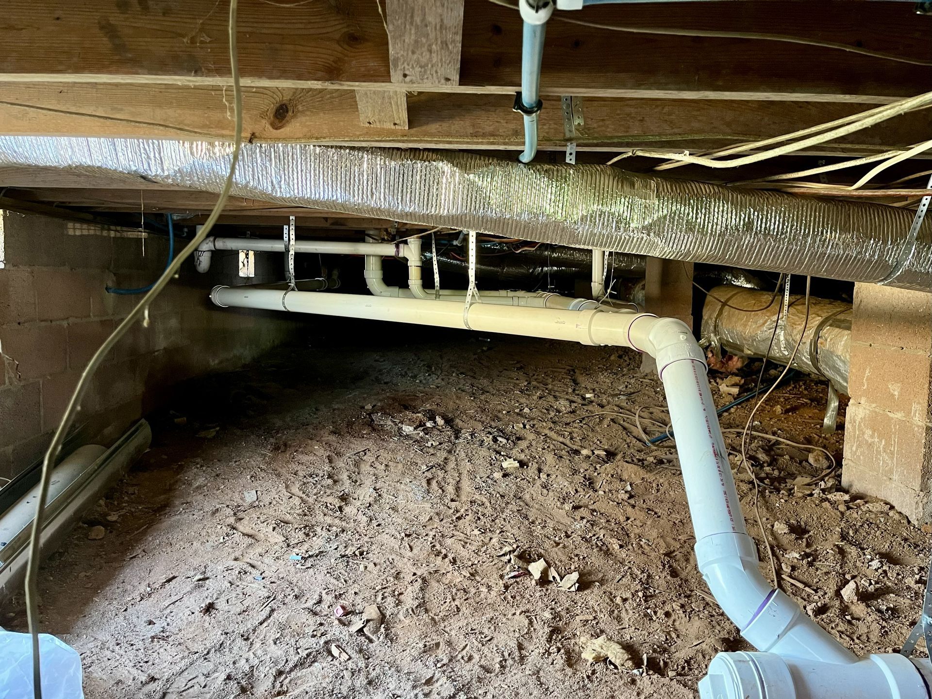 Underneath a house, PVC pipes, ductwork, and wiring run through a crawl space filled with dirt.