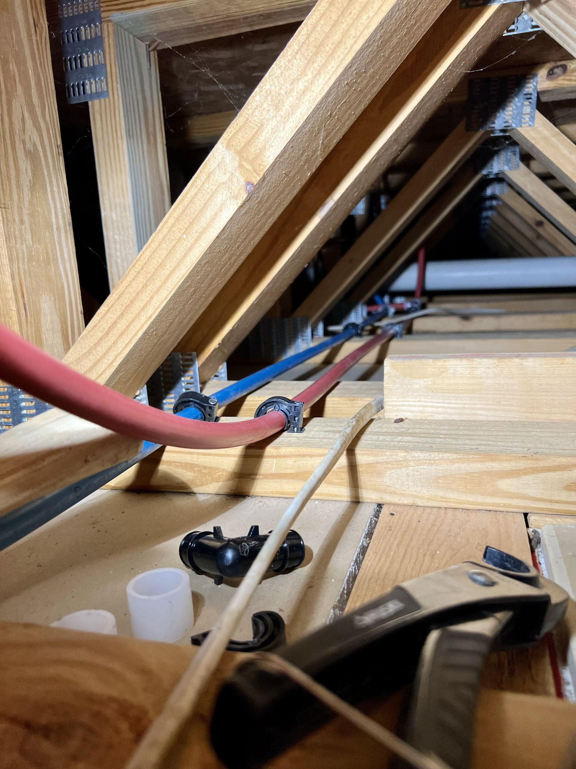 Red and blue water pipes secured to wooden rafters in an attic; various plumbing parts are scattered nearby.