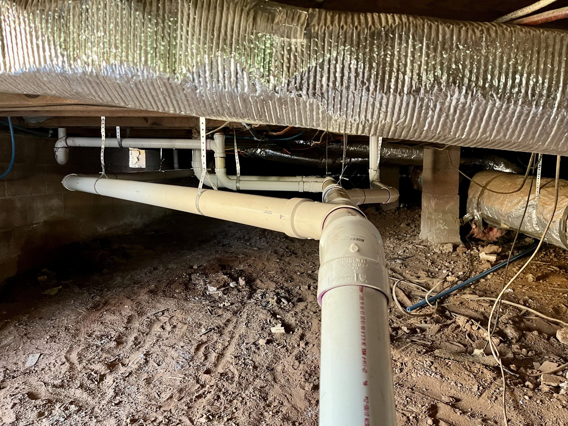PVC pipes and ductwork under a building. Dirt floor.