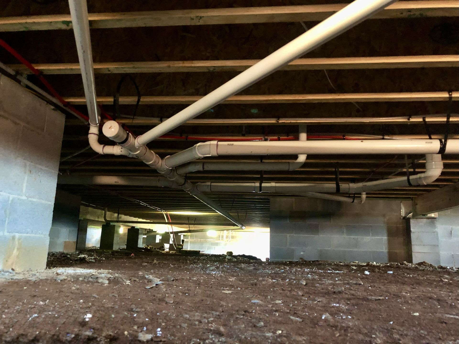 View of crawl space with white pipes and wooden beams. Ground is dirt, blocks support the structure.