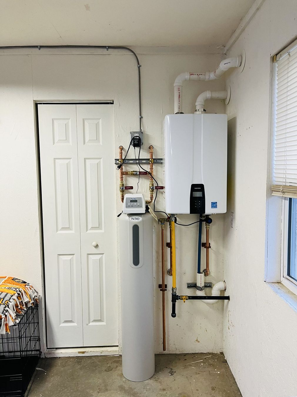 Water heater and water softener installed in a utility room near a door and window. White walls.