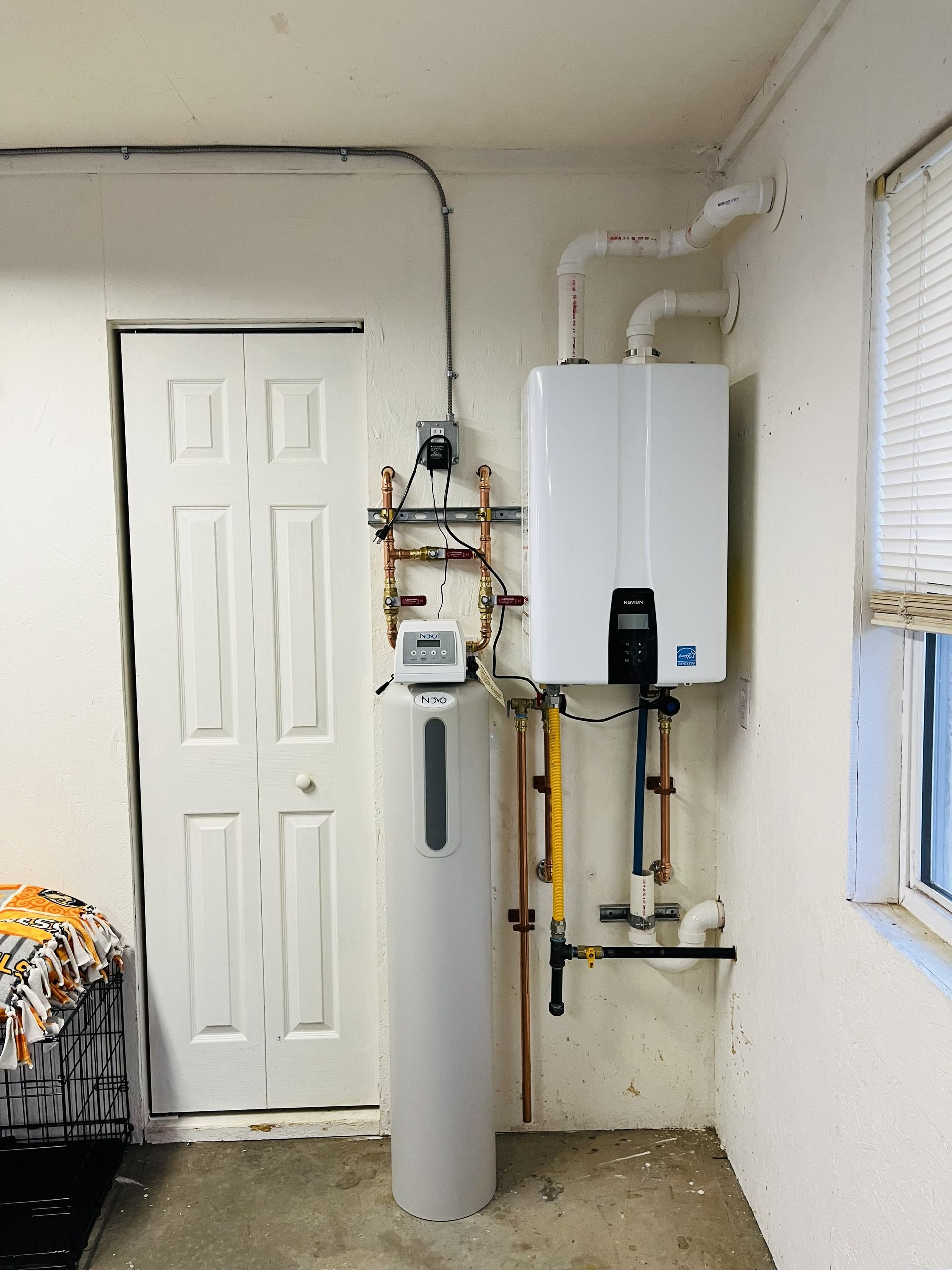 Water heater and water softener installed in a utility room near a door and window. White walls.