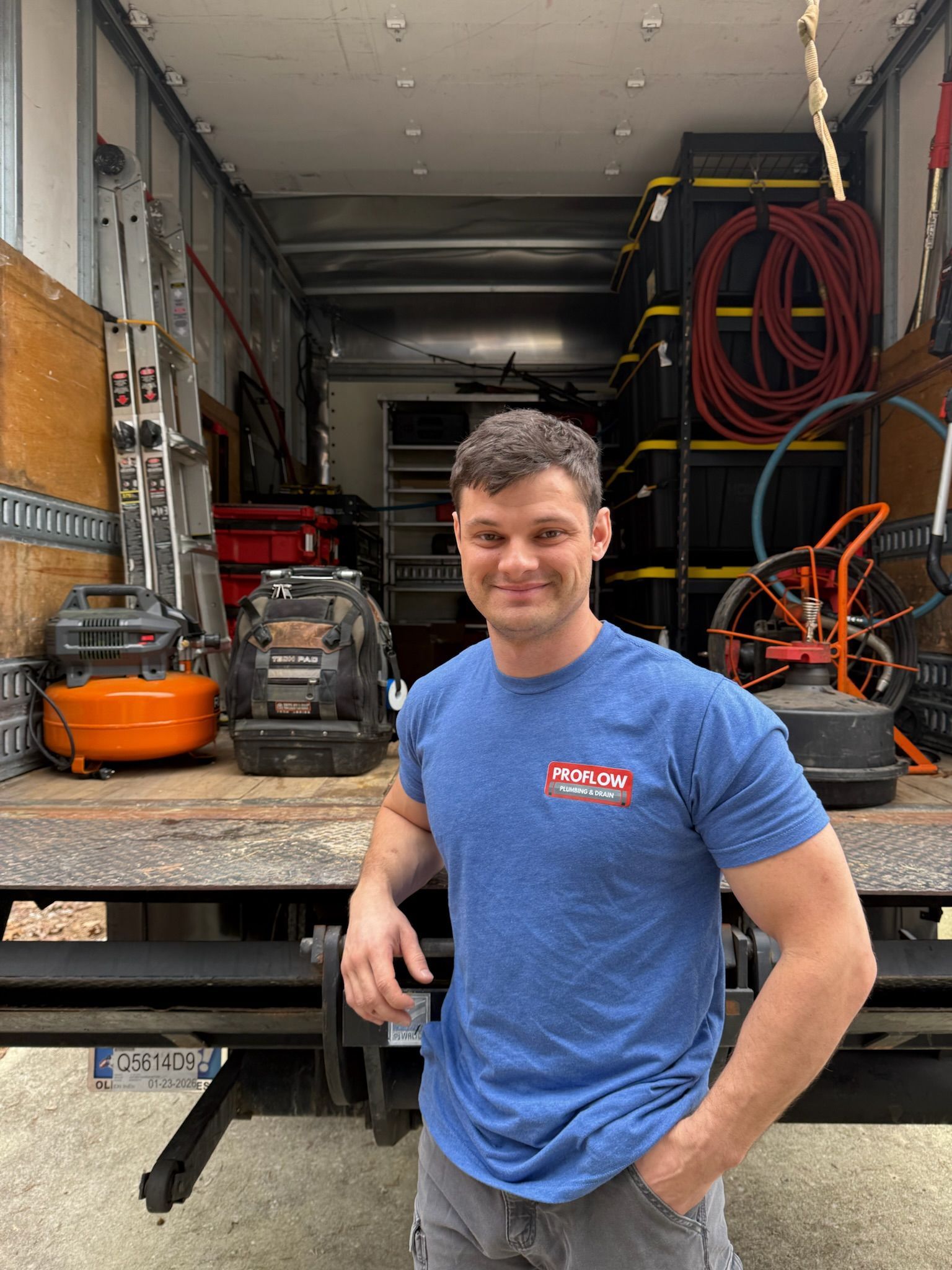 Man standing in front of a truck loaded with equipment, wearing a blue shirt. Smiling, with his hands in pockets.